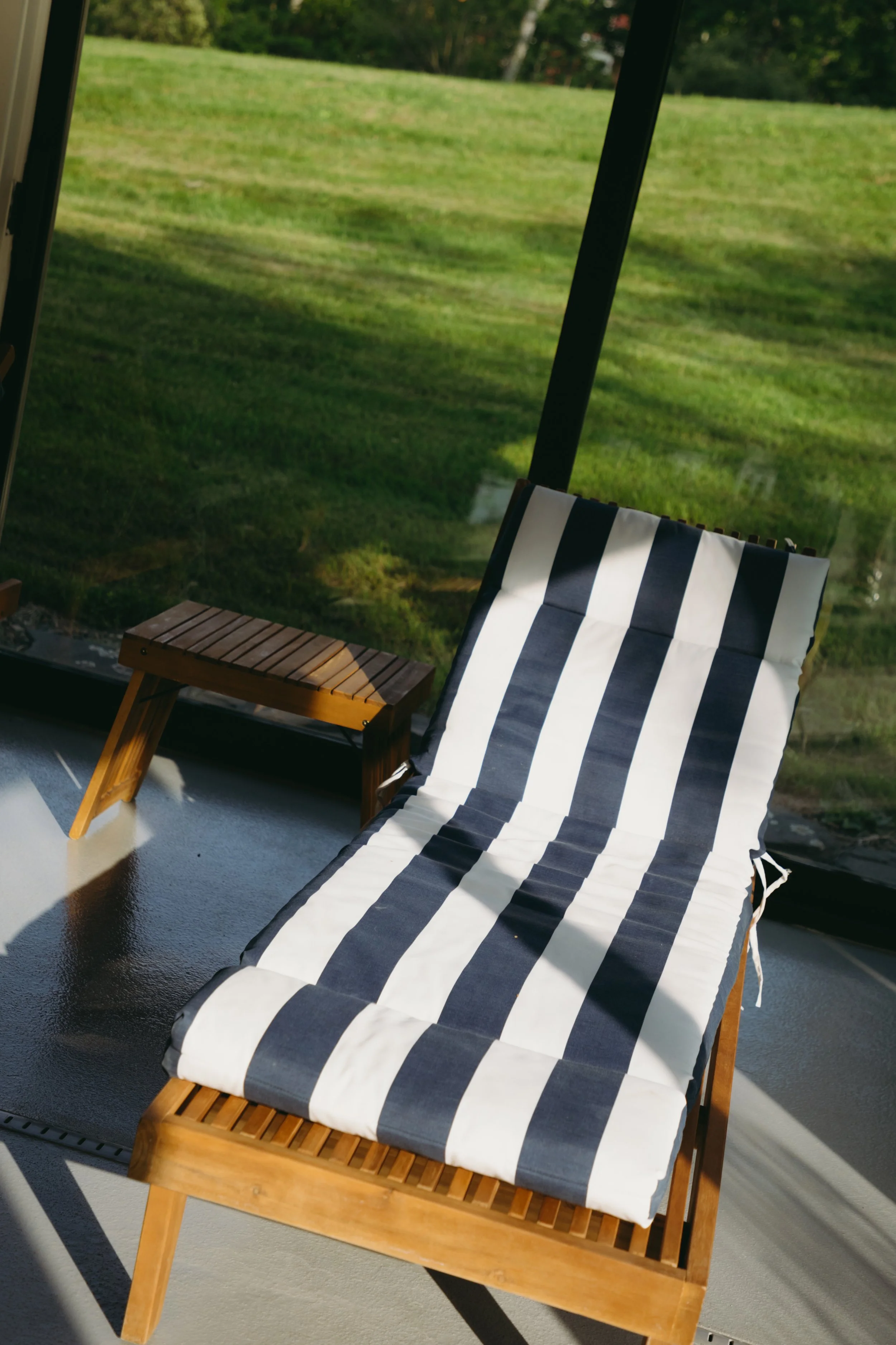 A wooden lounge chair with a black and white striped cushion on a patio or porch, with a view of green grass and trees outside.