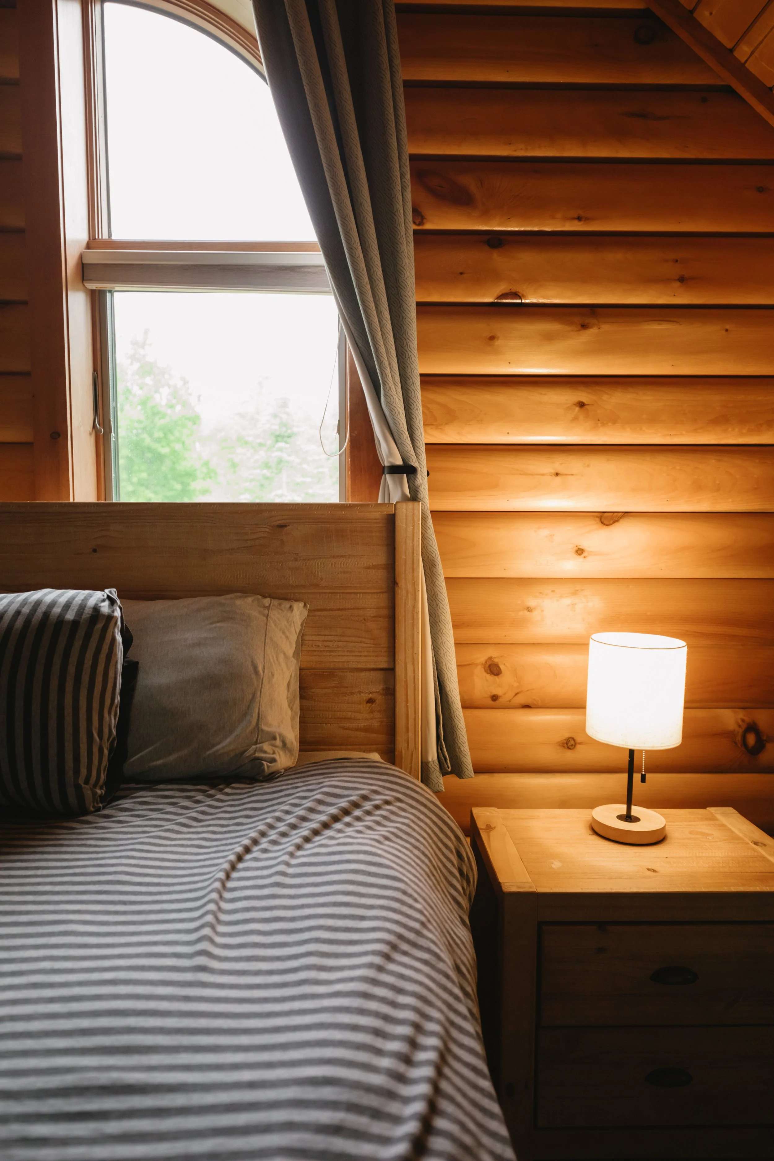 A cozy bedroom with wooden walls, a window with a curtain, a bed with striped sheets and pillows, and a table lamp on a wooden nightstand.