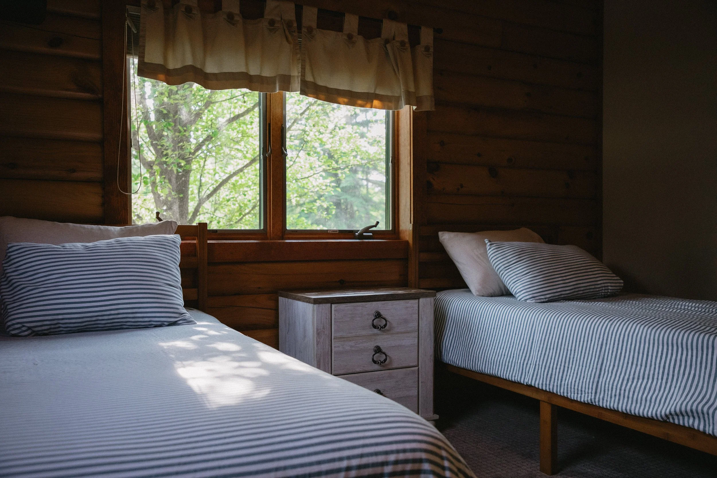 A cozy wooden cabin bedroom with two twin beds featuring blue and white striped bedding, a small white nightstand with black metal handles between the beds, and a window showing green trees outside.