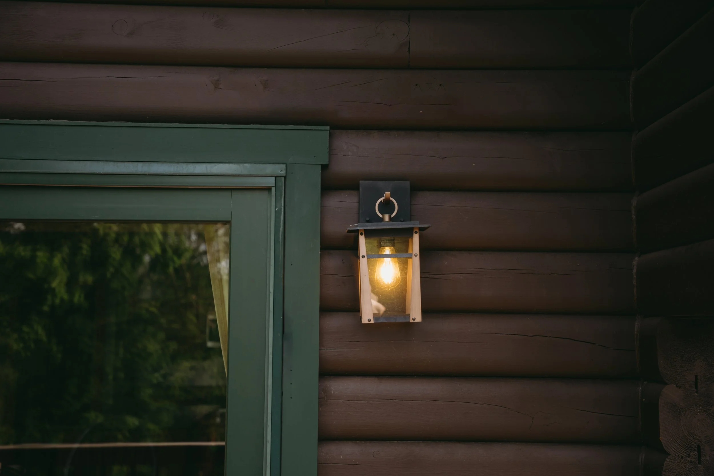 A wall-mounted outdoor lantern with a glowing bulb is attached to a dark wooden log cabin wall next to a window with a green frame.