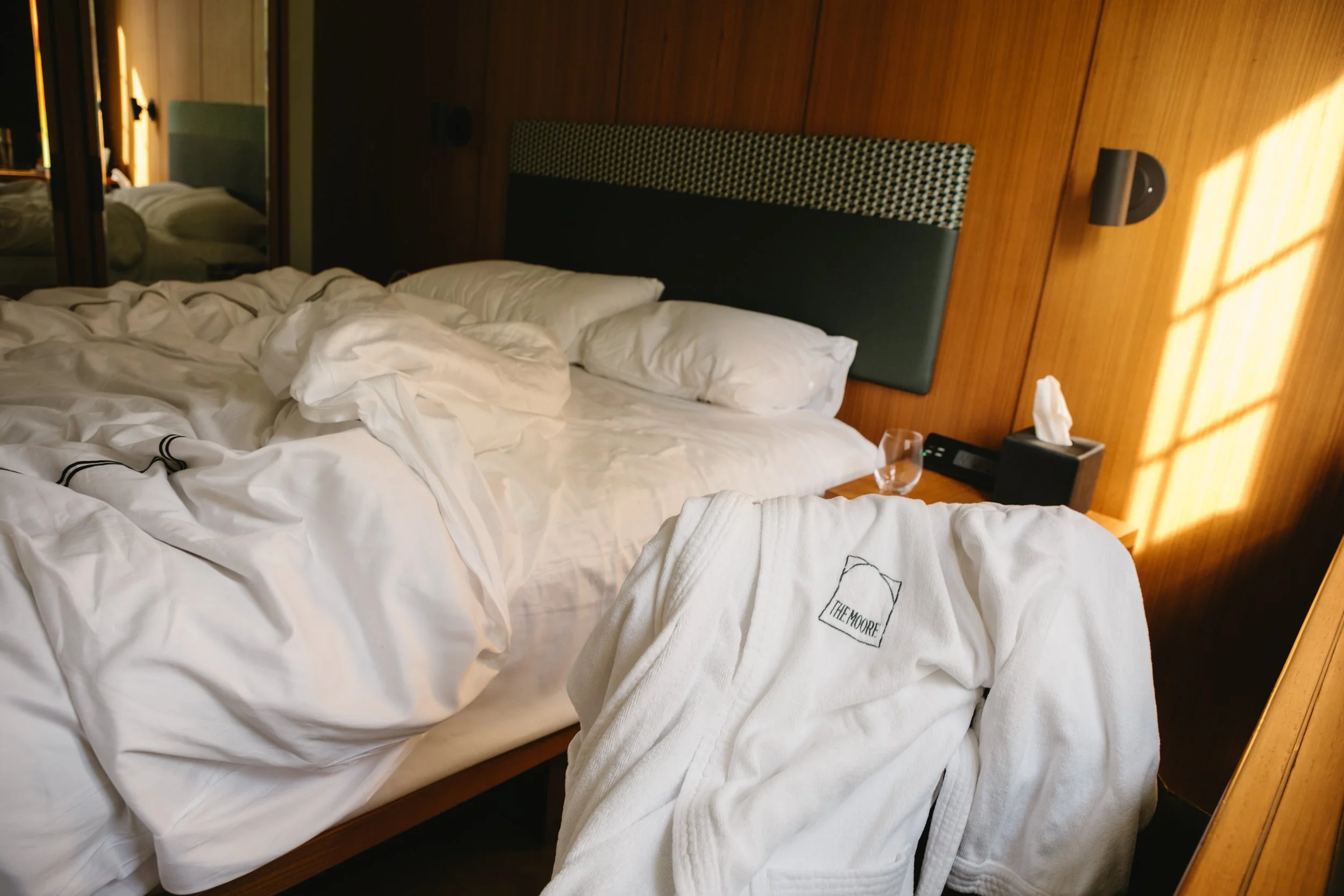 A messy hotel bed with white sheets and pillows, a robe embroidered with 'The Moore', a glass of water, a tissue box, and a wooden headboard in a room with warm lighting.