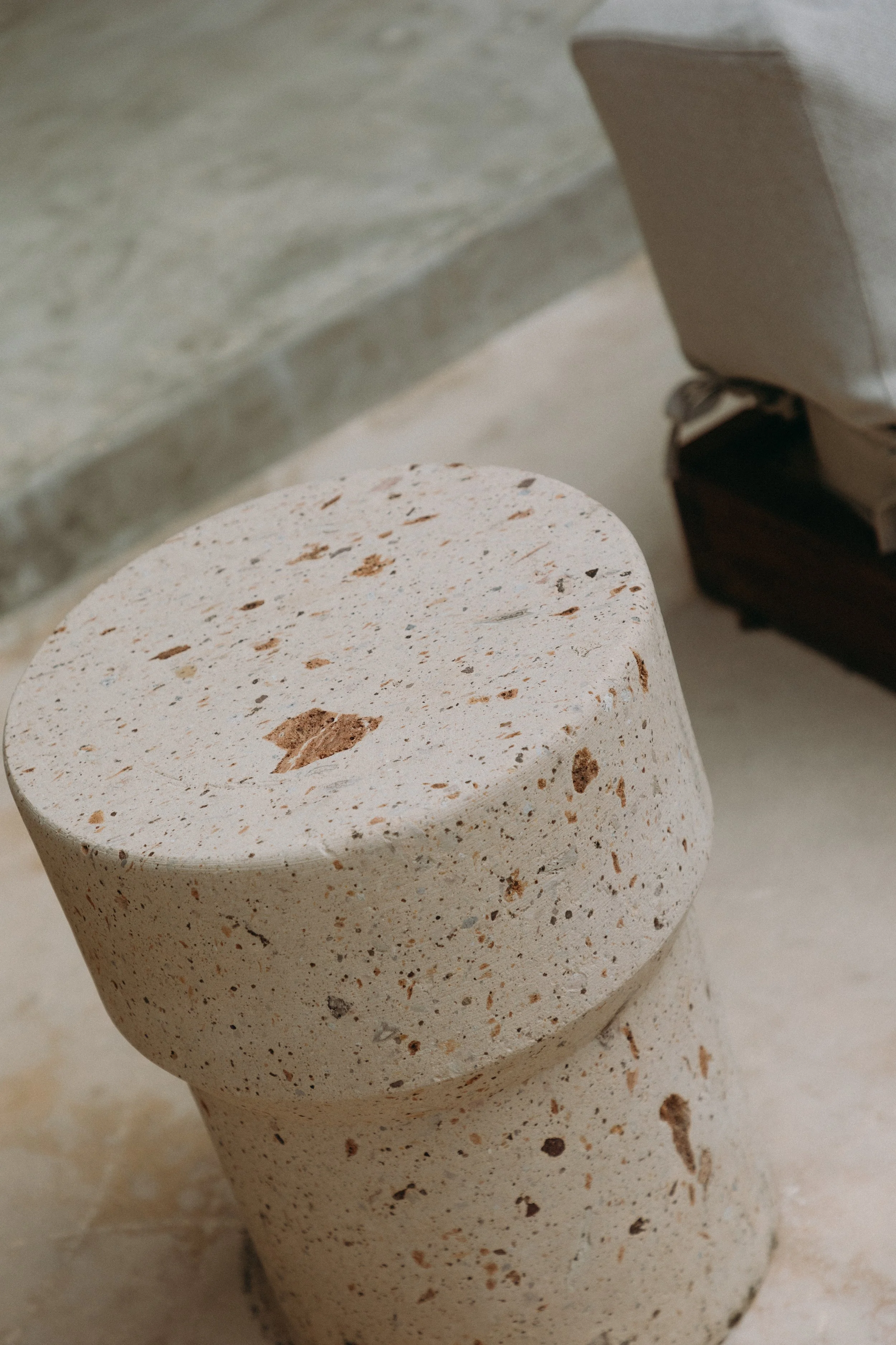 Close-up of a rounded, speckled stone or terrazzo-style stool or table with beige and brown tones, next to a sofa or chair with a brown wooden frame and light-colored cushion.