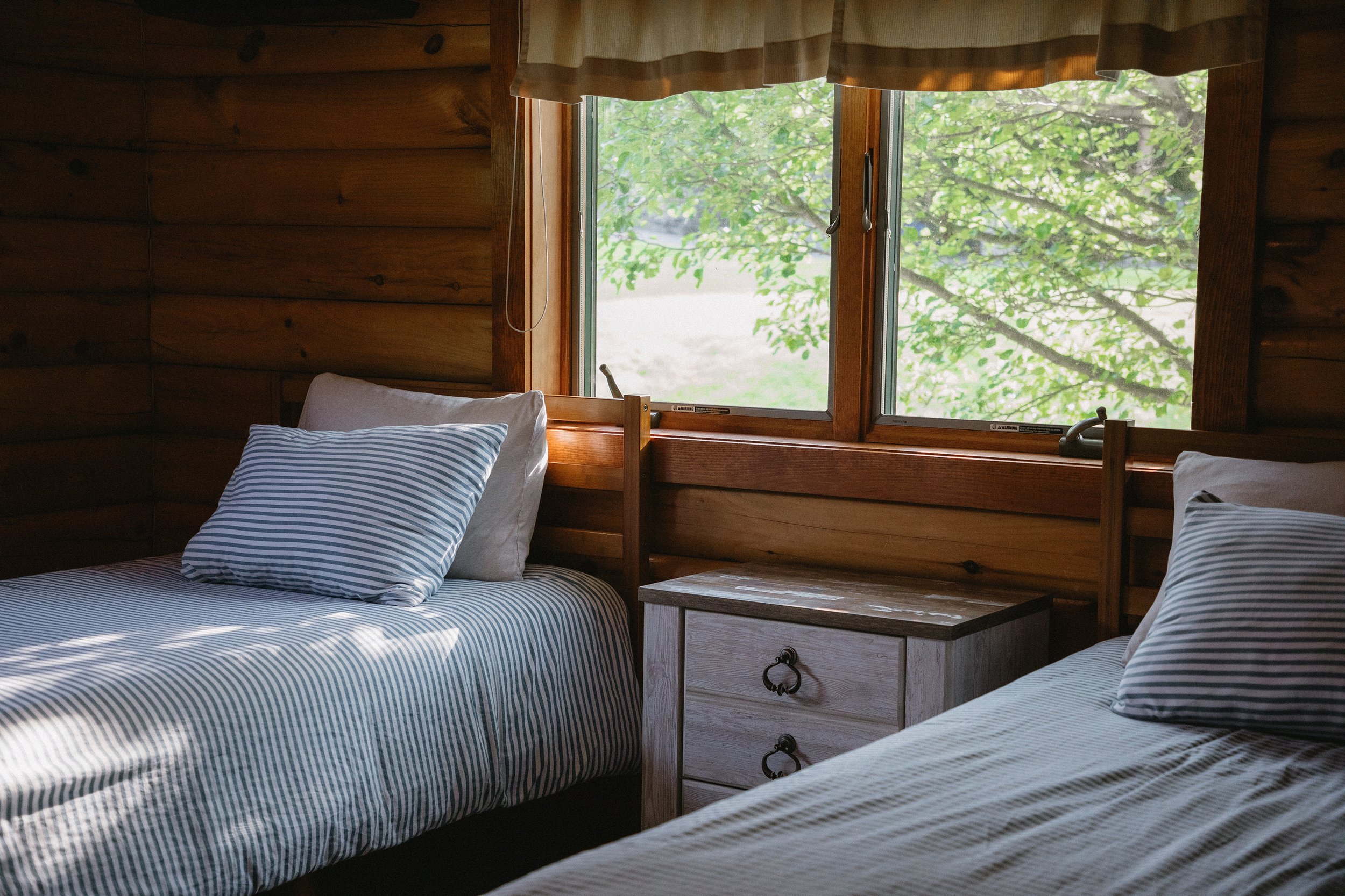 Two twin beds with striped bedding and pillows in a wooden cabin bedroom, with a window showing green trees outside.