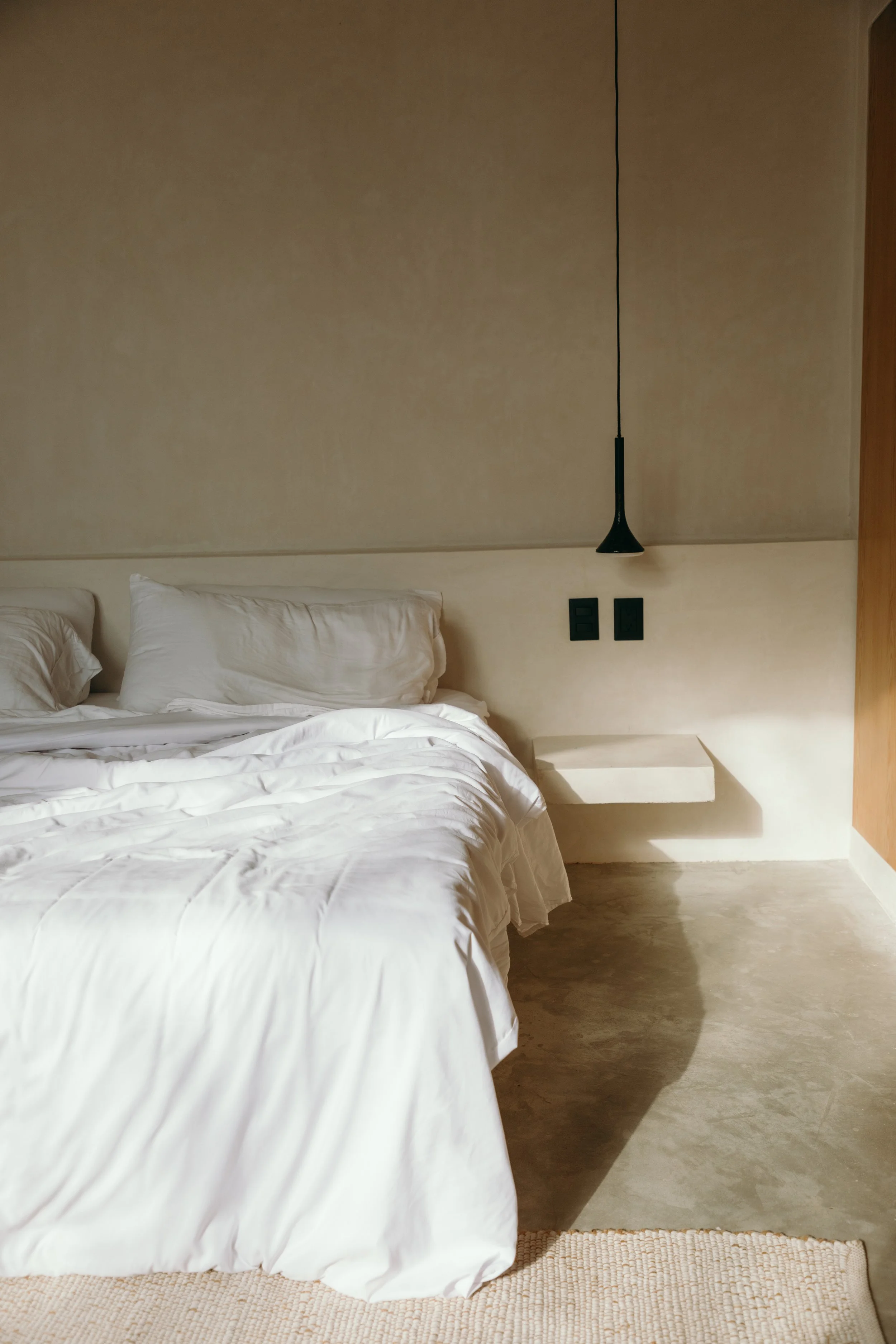 A bedroom with a neatly made bed with white linens and pillows, a small floating nightstand, a hanging black pendant light, and a concrete floor with a beige rug.
