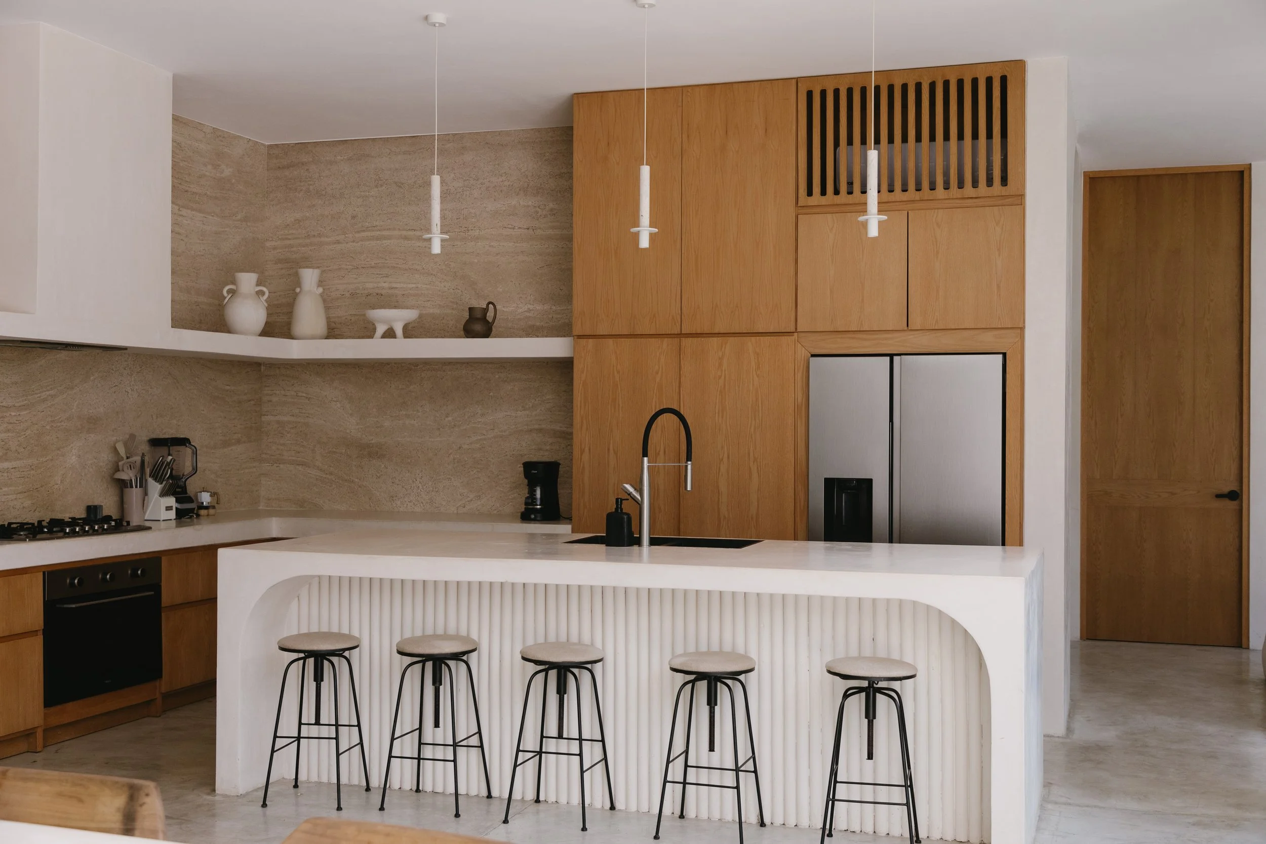 Modern kitchen with wooden cabinets, white island with a ribbed design, black sink and faucet, barstools, stainless steel refrigerator, and minimalist decor.