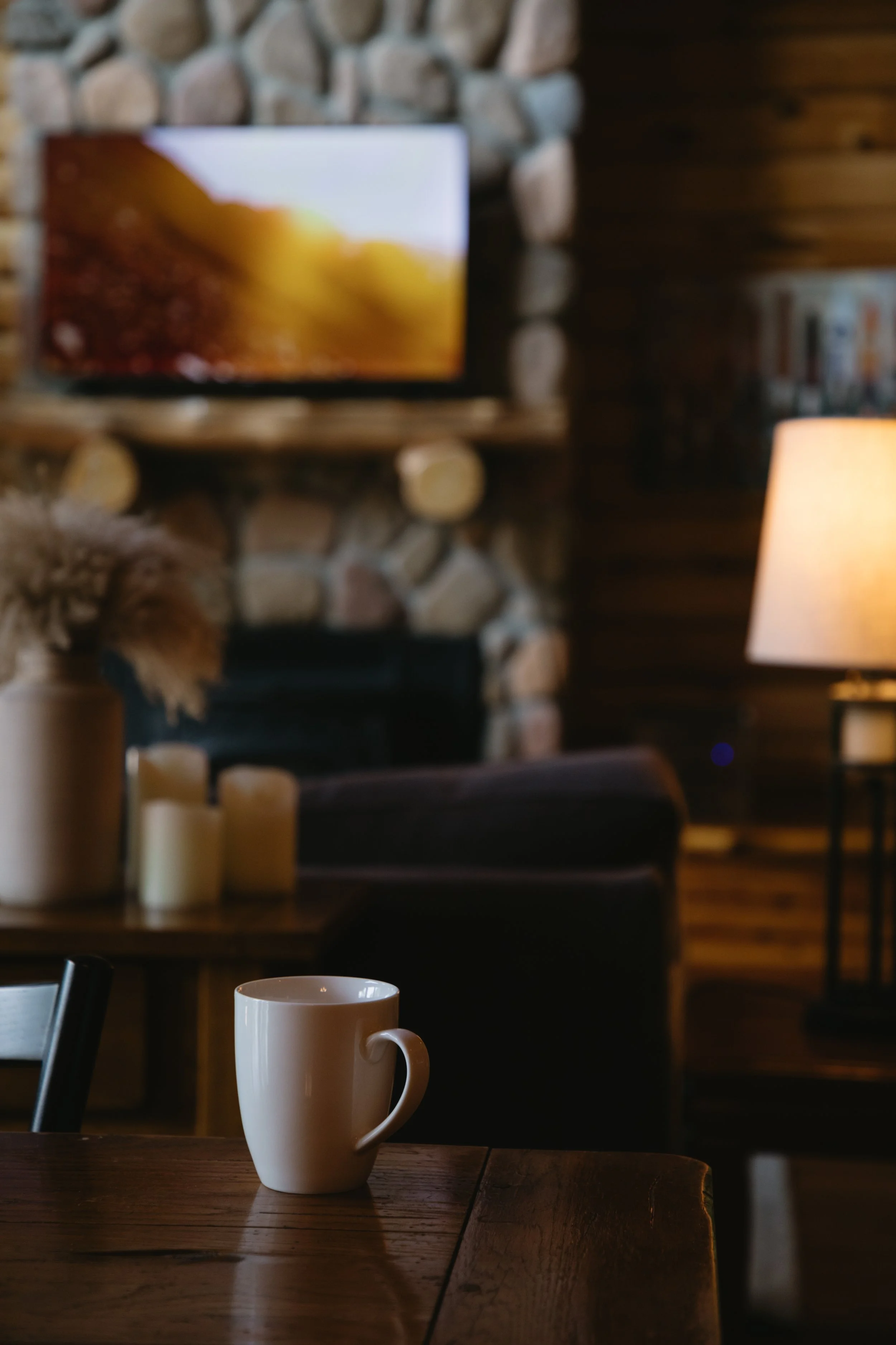 A white coffee mug on a wooden table in a cozy living room with a stone fireplace, a lamp, candles, and a television mounted on the wall.
