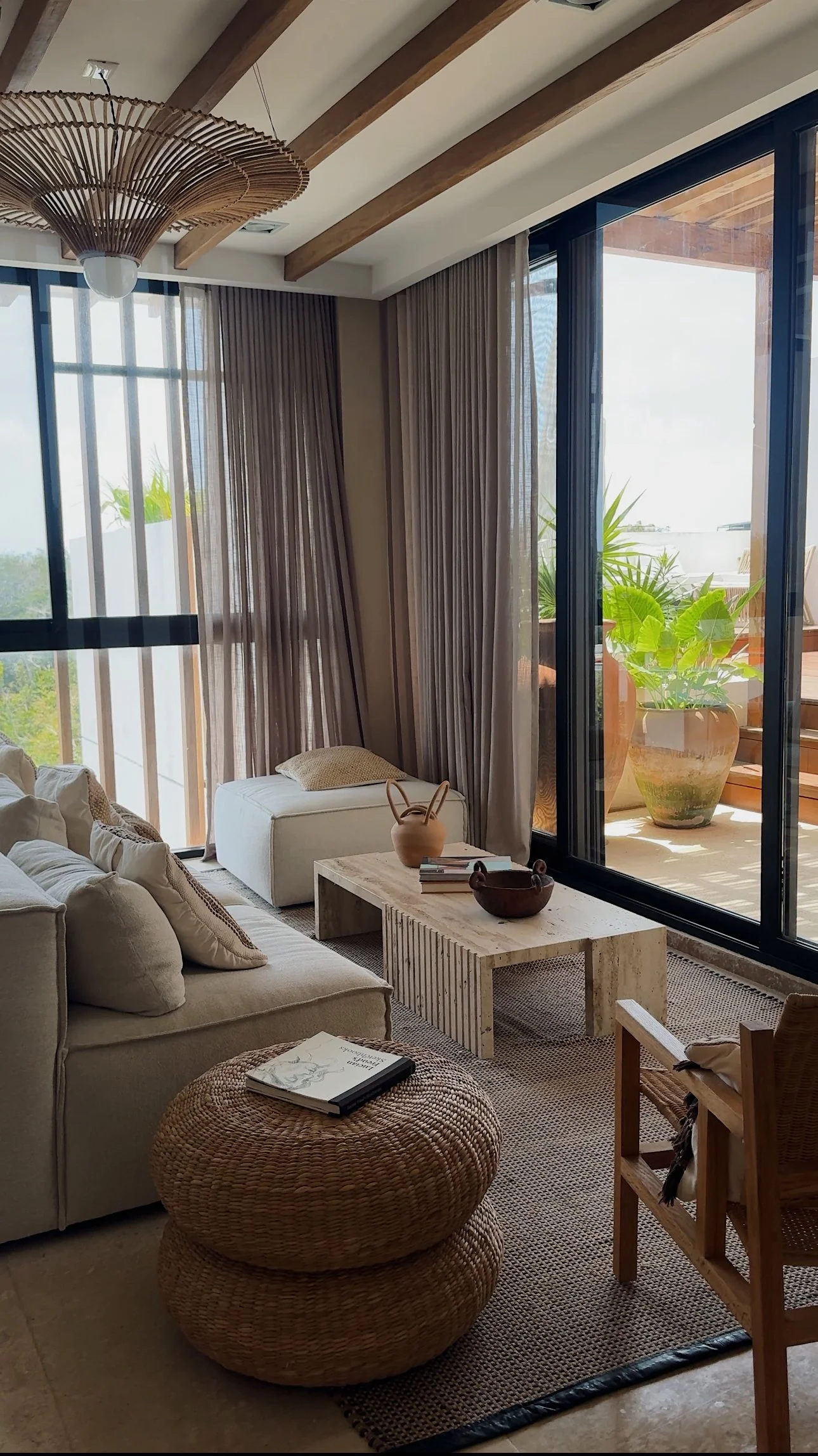 A cozy living room with beige sofa, wicker pouf, and wooden coffee table. Large glass sliding doors lead to a balcony with potted plants, letting in natural light.