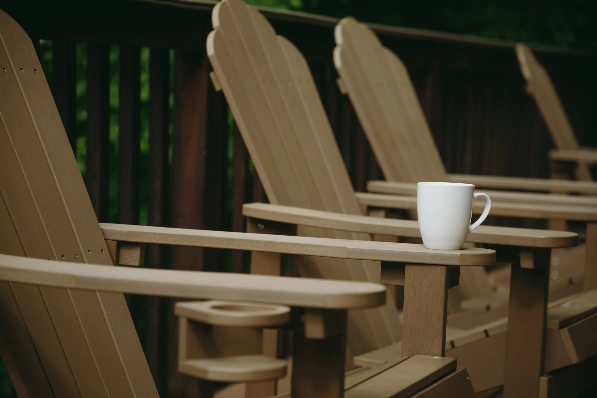 A row of wooden Adirondack chairs outdoors with a white mug placed on the armrest of the front chair.