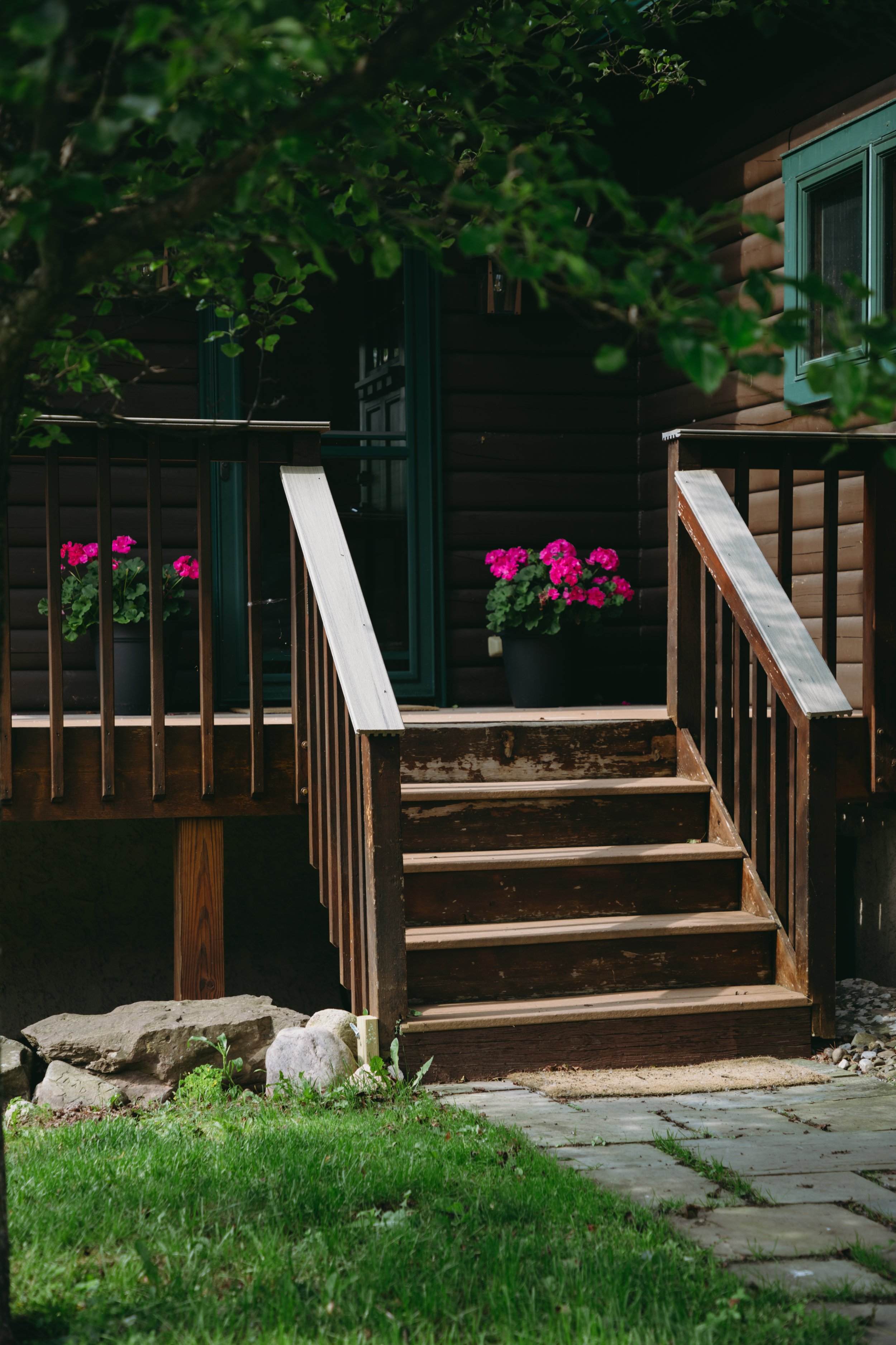 Wooden deck and stairs leading to a door of a house with dark siding, pink flowers in pots, and a small stone border.
