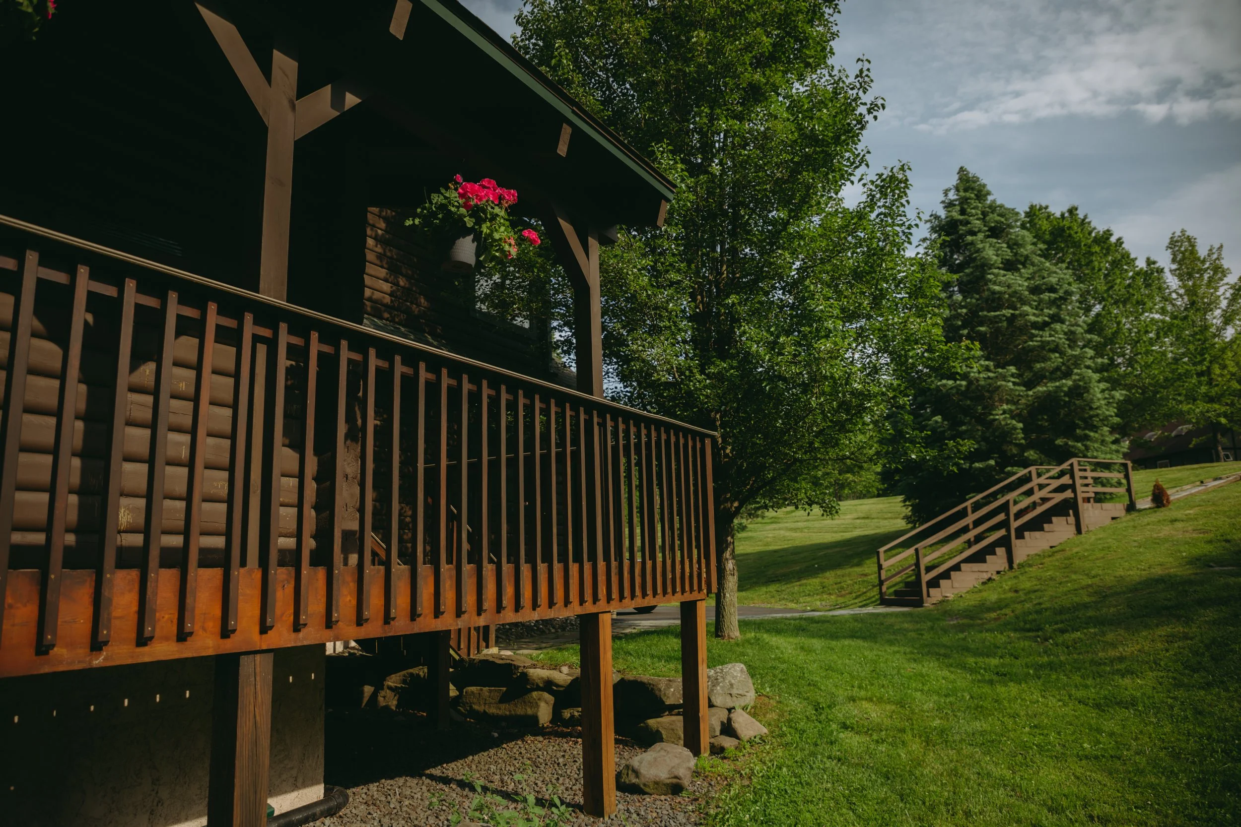 A wooden deck with a dark railing attached to a house, overlooking a grassy yard with stairs leading up a small hill, surrounded by green trees under a partly cloudy sky.