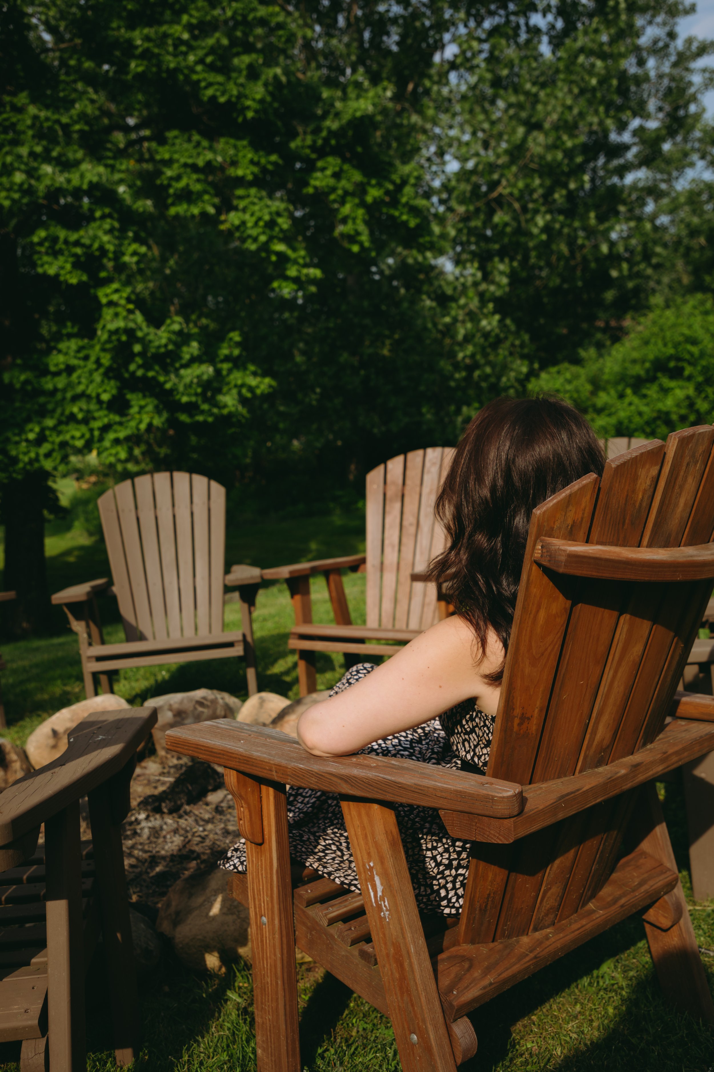 A woman with dark hair sitting in a wooden Adirondack chair outdoors with lush green trees in the background.