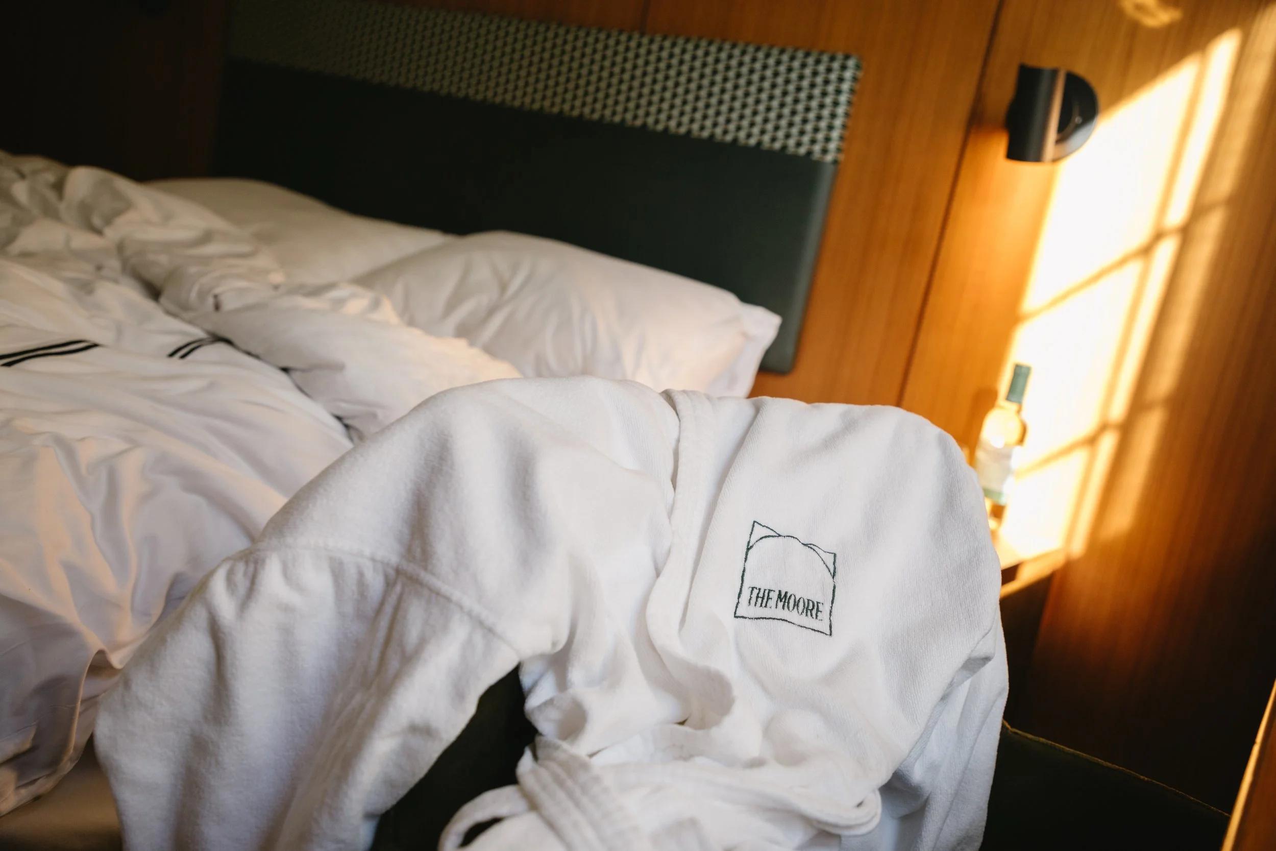 Unmade bed with white sheets and pillows, a white bathrobe with 'The Moore' embroidered on it, on a dark surface, and a wooden wall with a lamp and a bottle on a bedside table in the background.