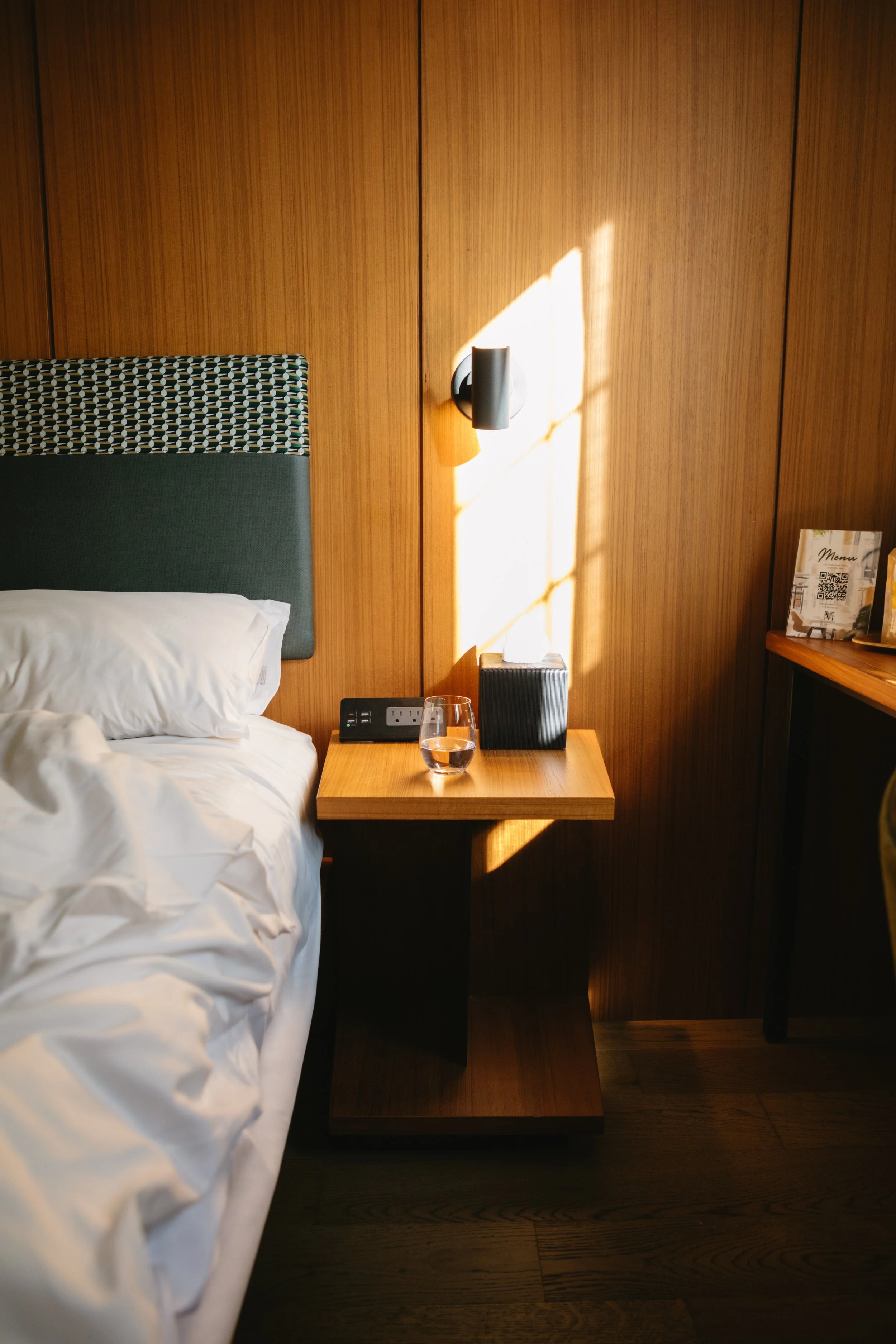 A hotel room with a bed, a wooden nightstand with a glass of water, a tissue box, and a wall-mounted light, illuminated by sunlight through a window.