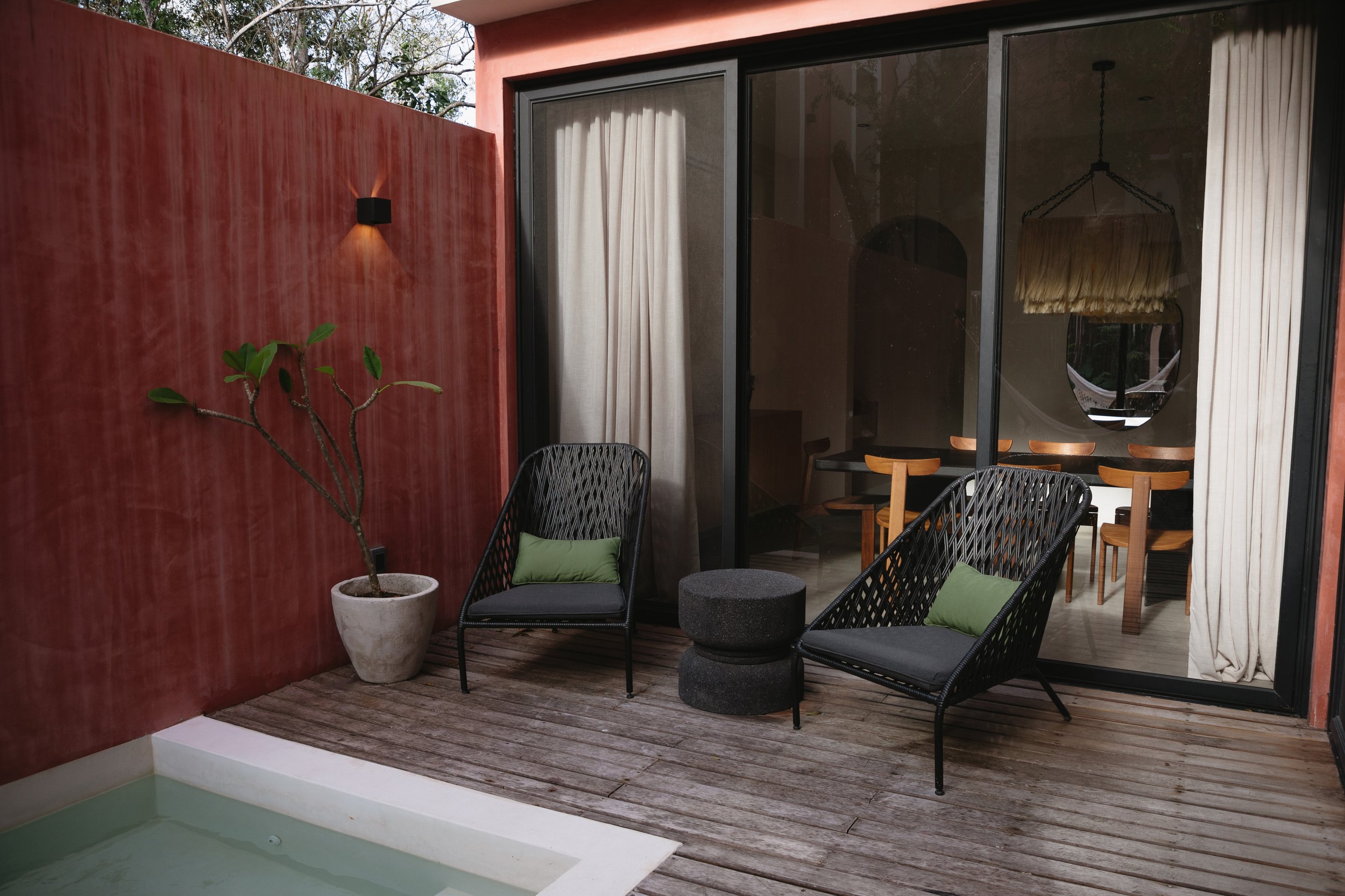 A small outdoor patio area with two black woven chairs, green cushions, a small round black side table, and a potted plant with a twisted trunk. There is a red wall with a wall-mounted light fixture, and a sliding glass door revealing an indoor dinin