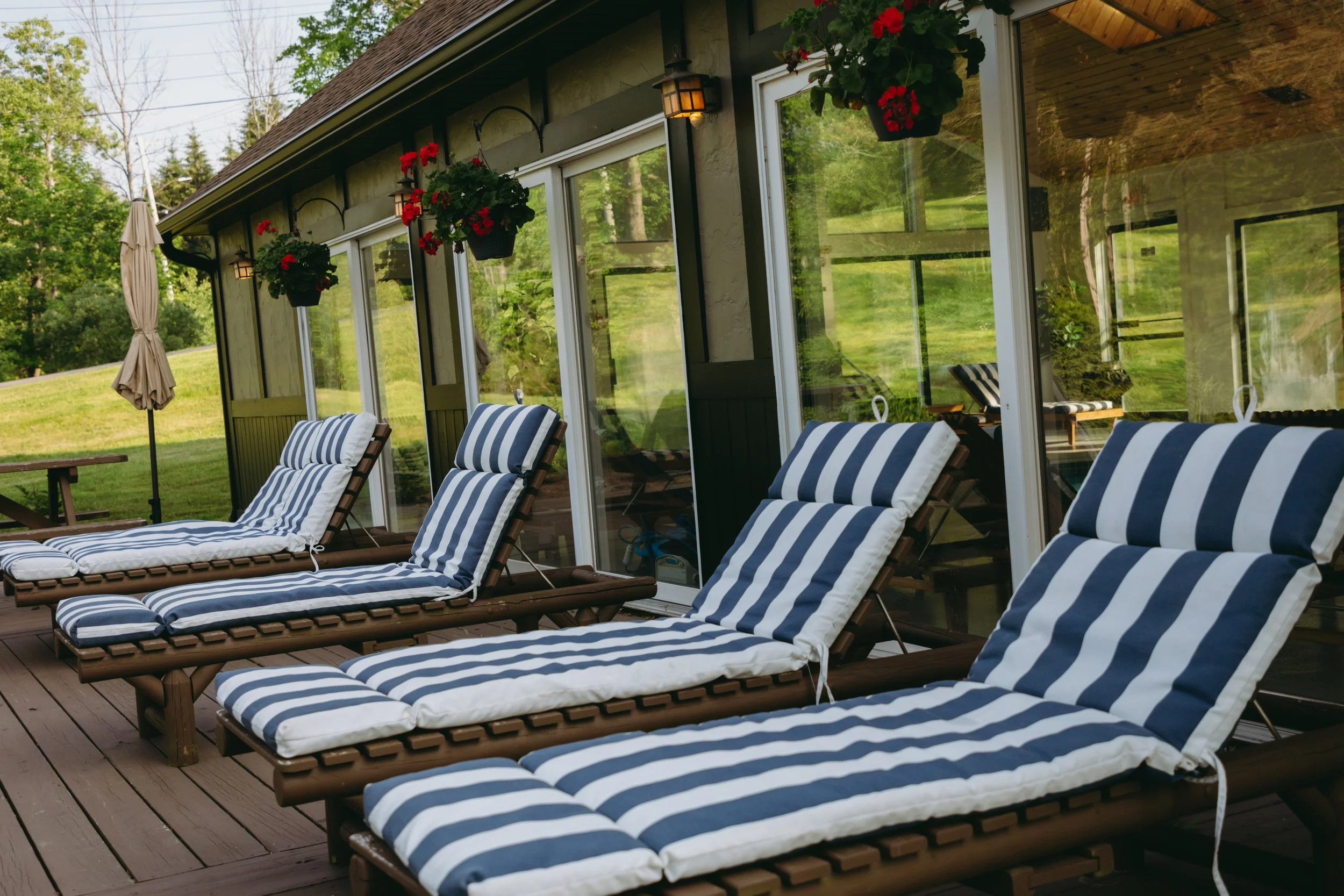 Chairs with striped cushions on a wooden deck outside a house with large glass sliding doors, decorated with hanging flower pots and outdoor lights, overlooking a grassy yard.