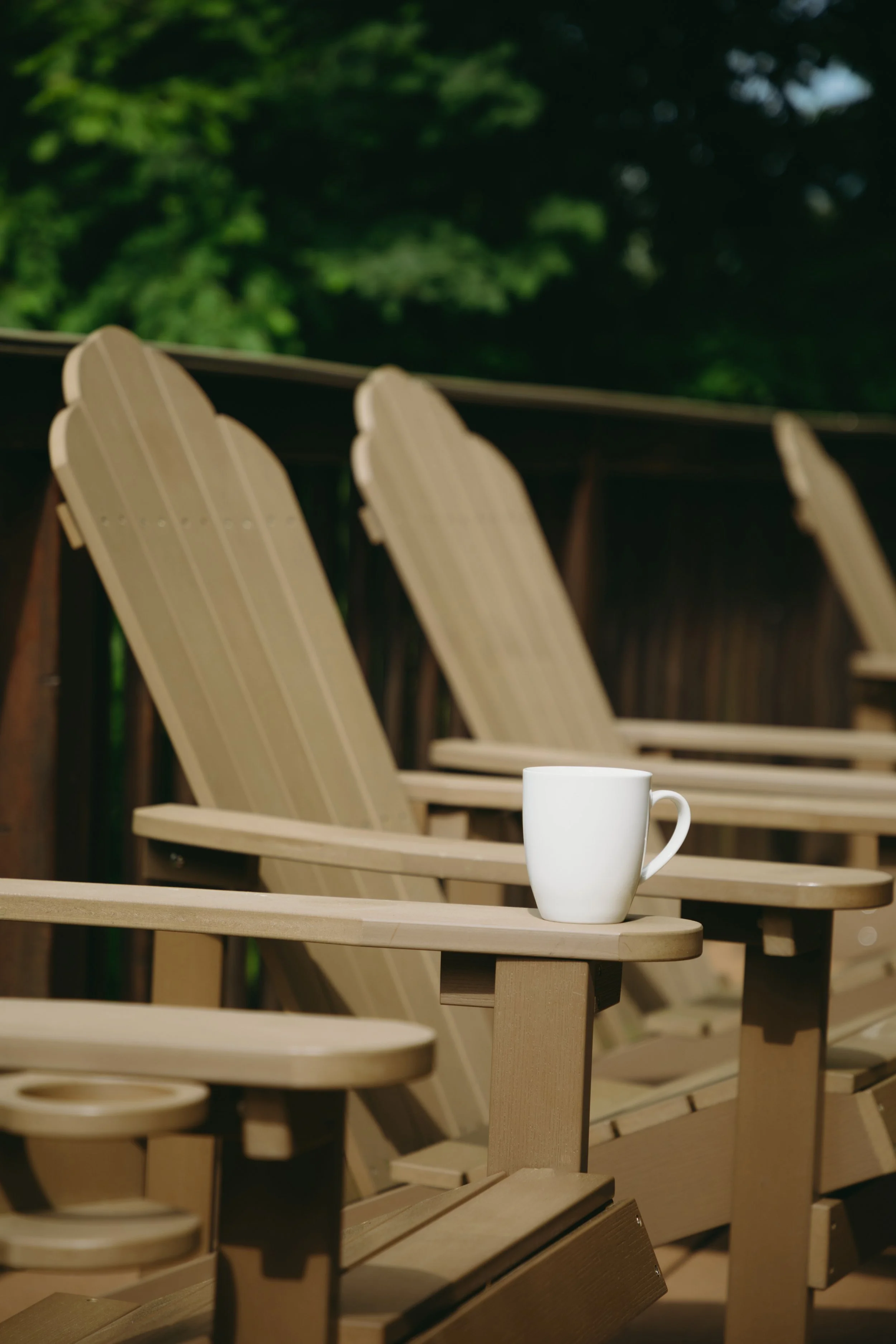 A white mug placed on an outdoor wooden armrest of a tan Adirondack chair, with more chairs in the background and green foliage in the distance.