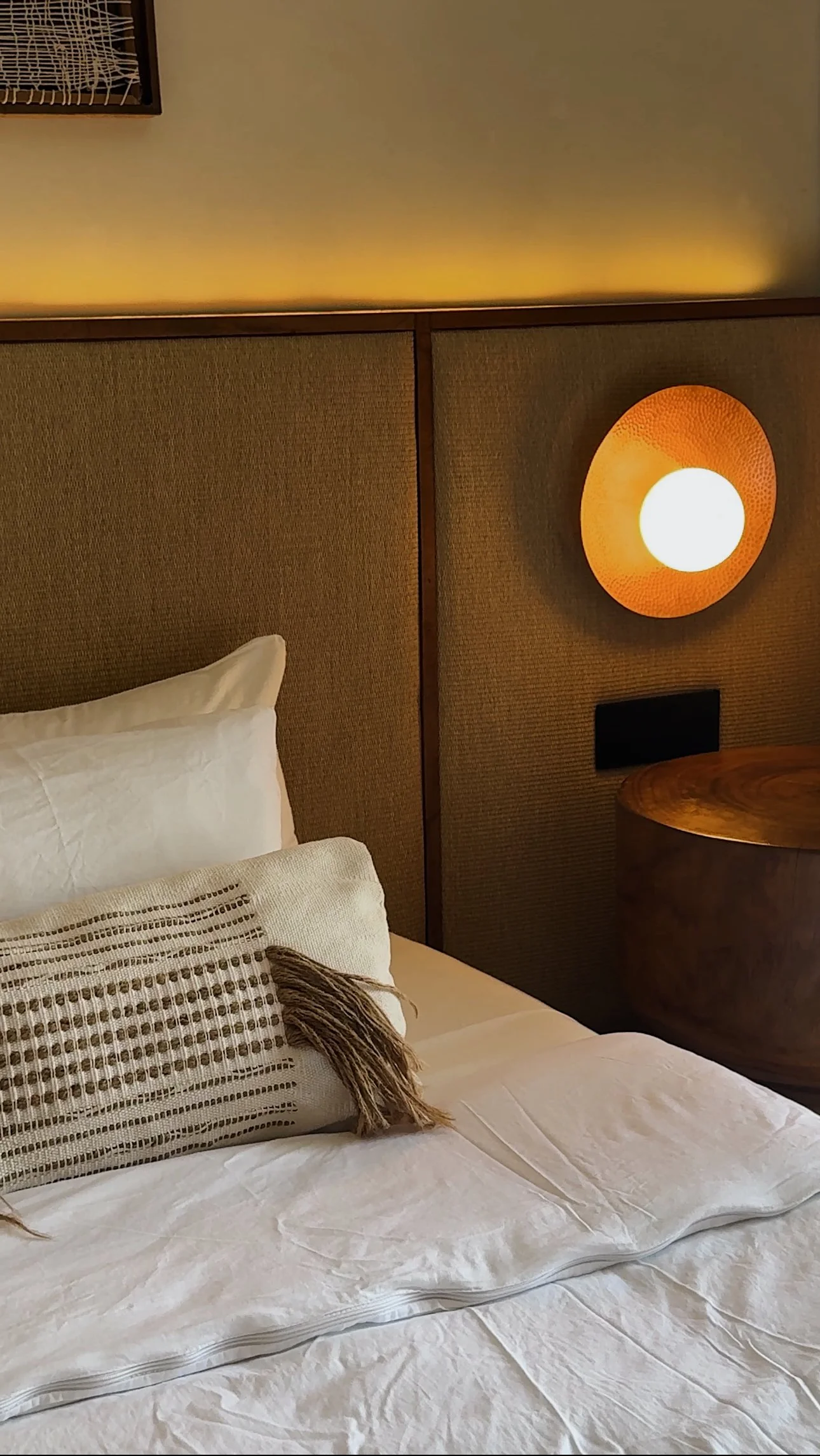 Close-up of a neatly made bed with white linens, decorative pillows, and a woven throw, in a hotel room with a textured brown wall, wood accents, and a round orange wall sconce.