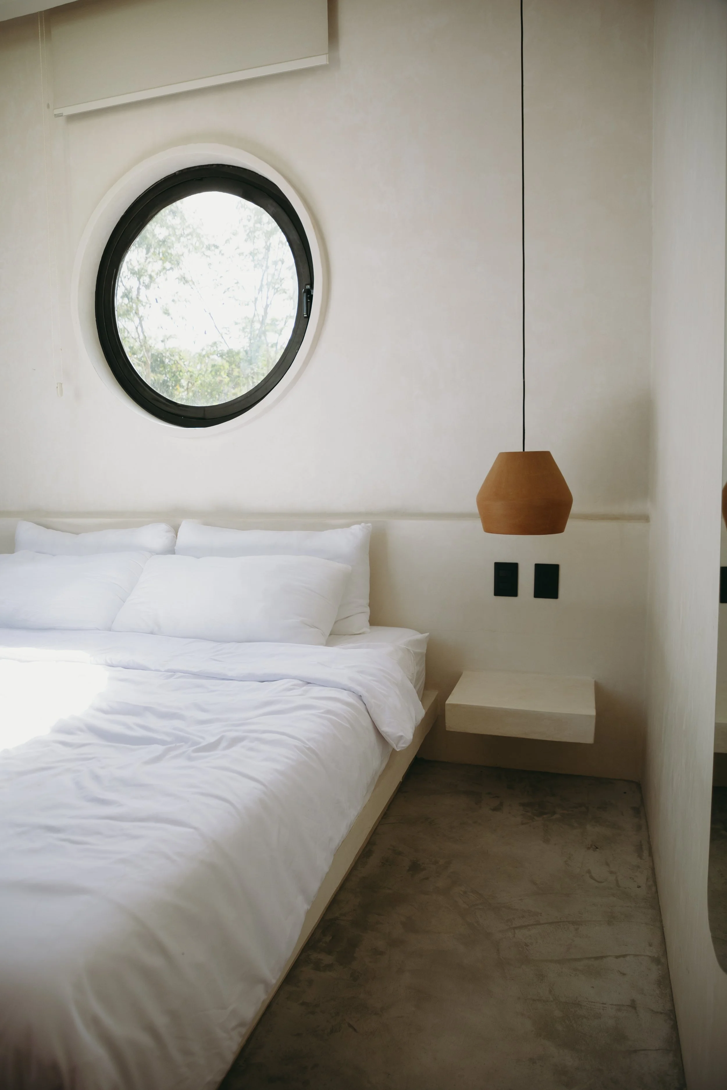 A minimalist bedroom with a round window, white bed with pillows, a hanging wooden lamp, and a small floating nightstand against light-colored walls.