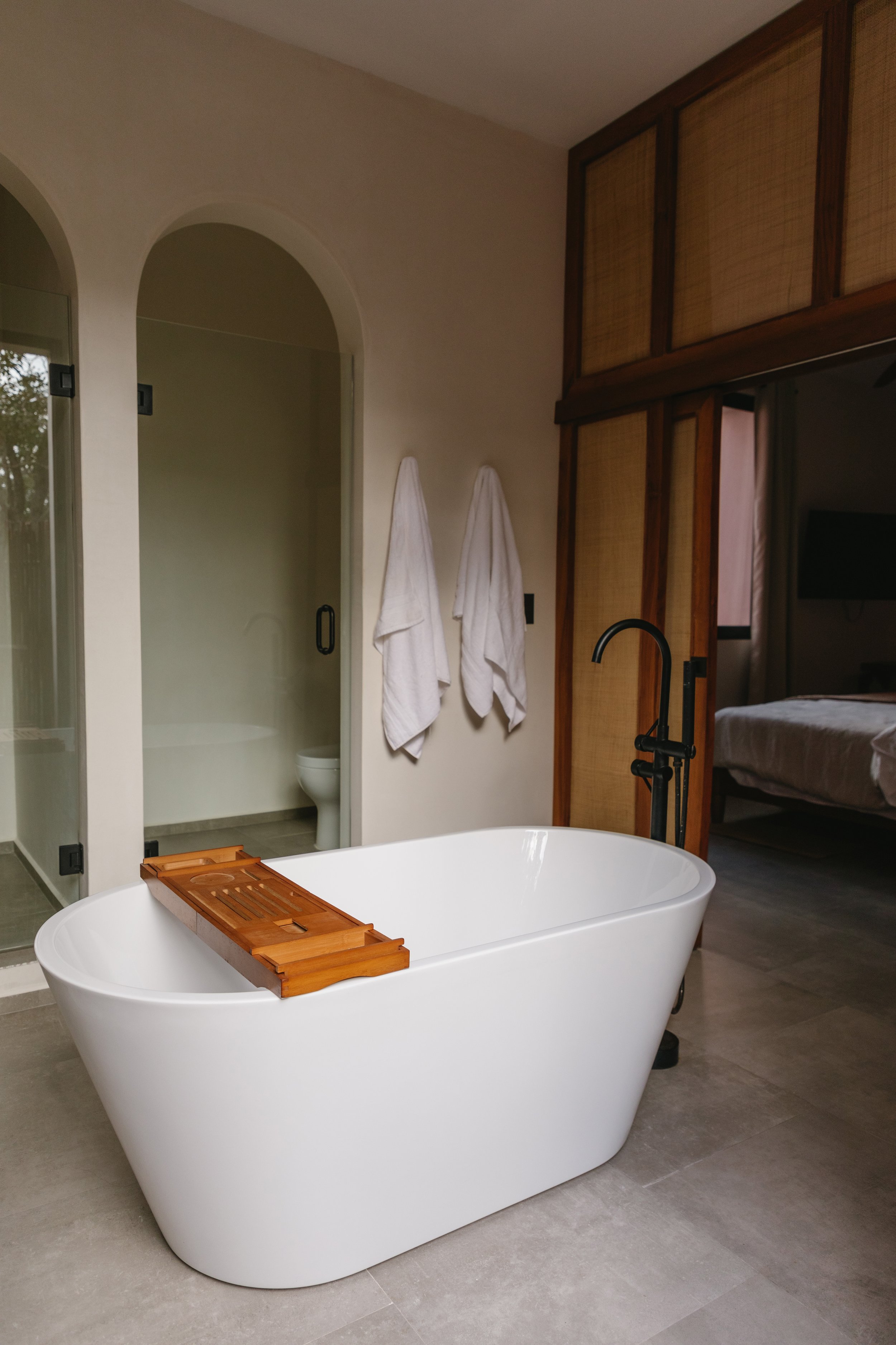 Modern bathroom with a freestanding bathtub, wooden tray, towel hooks, and an adjacent open bedroom with a bed.