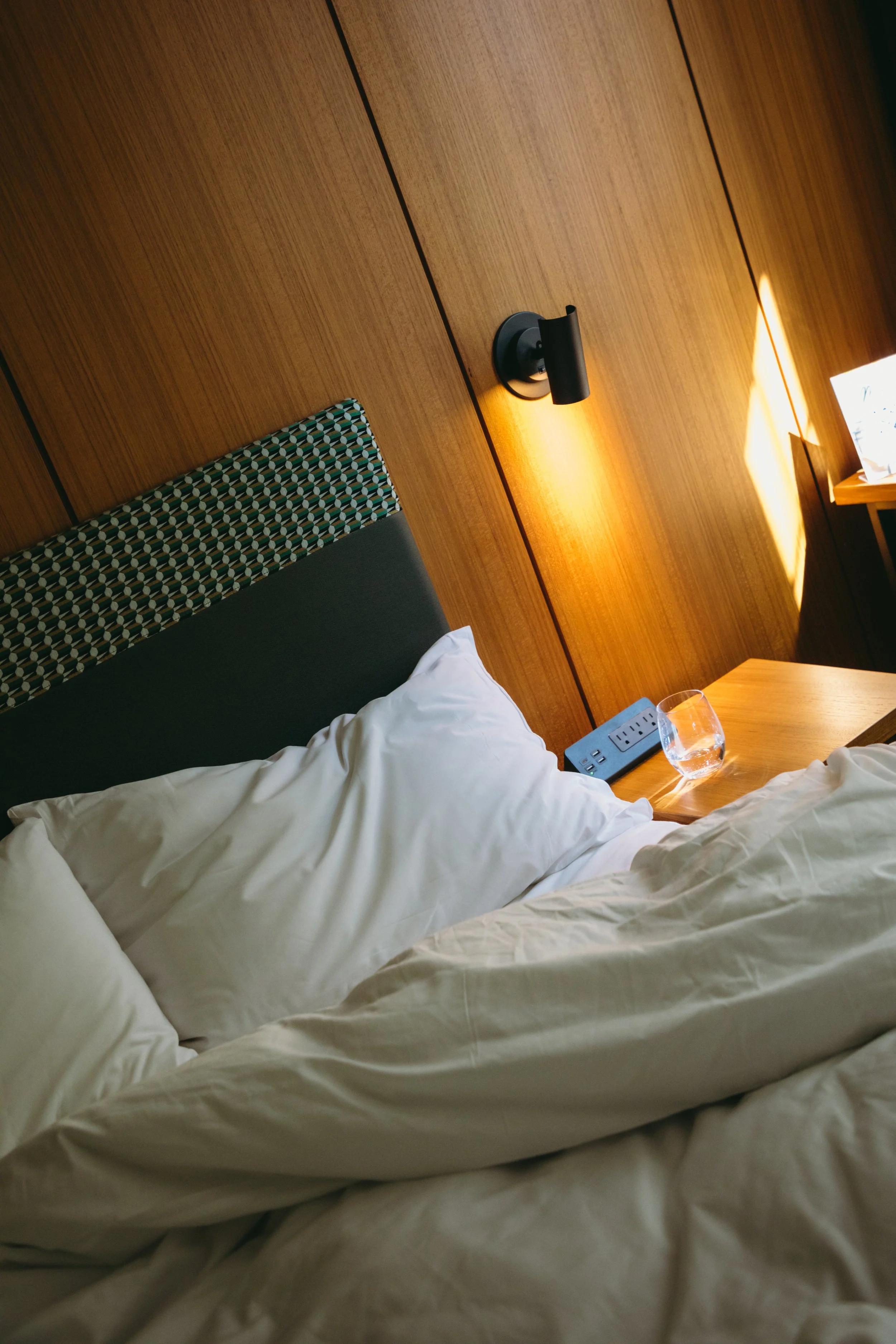 Unmade bed with white pillows and sheets in a hotel room, with a wooden headboard, nightstand, glass of water, and a wall-mounted lamp.