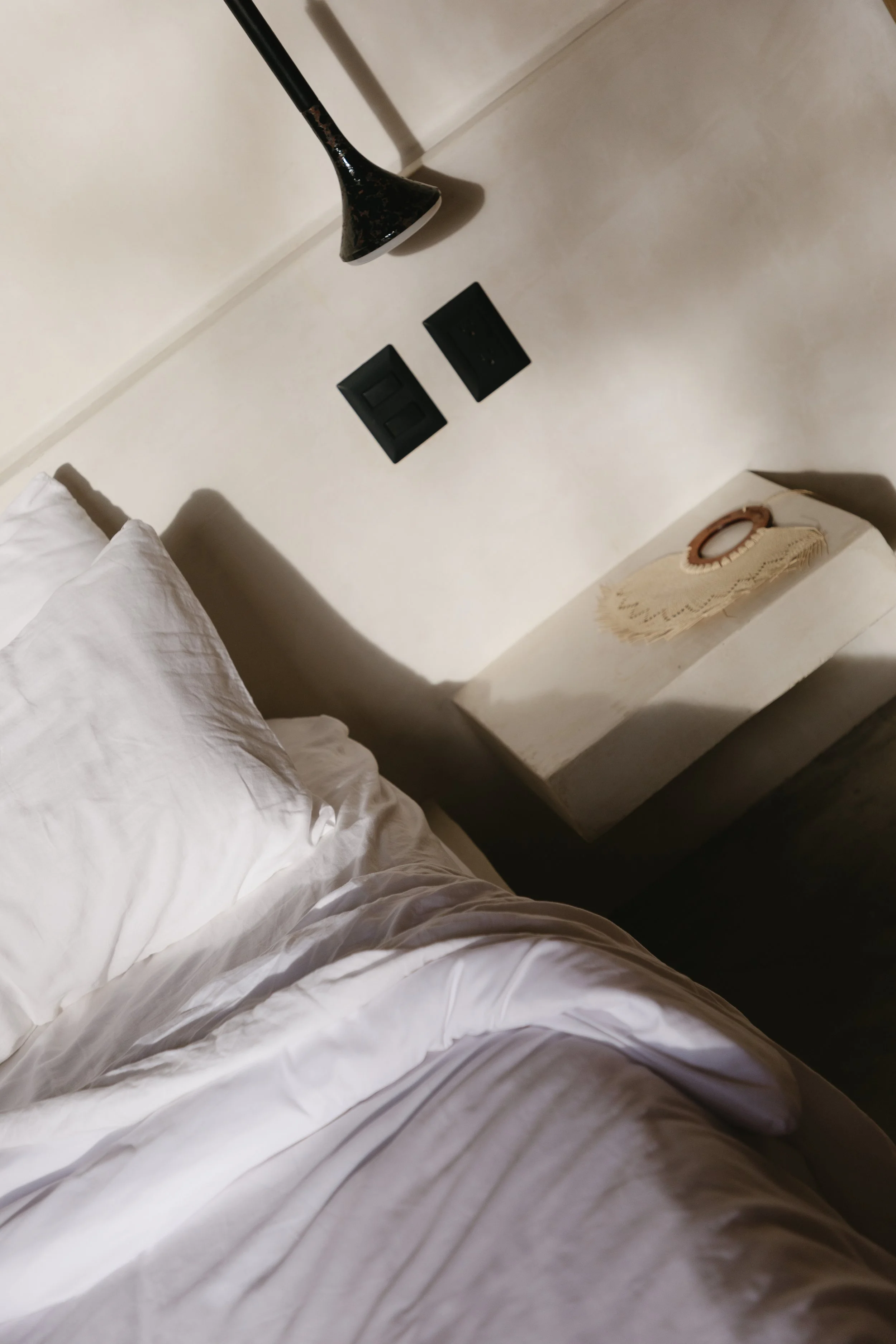 Part of a crispy white bed with white pillow and sheets, above a white nightstand with crocheted doily and small object, beige wall with two black electrical switches, white electrical outlet, and a black lamp with a cone-shaped shade nearby.