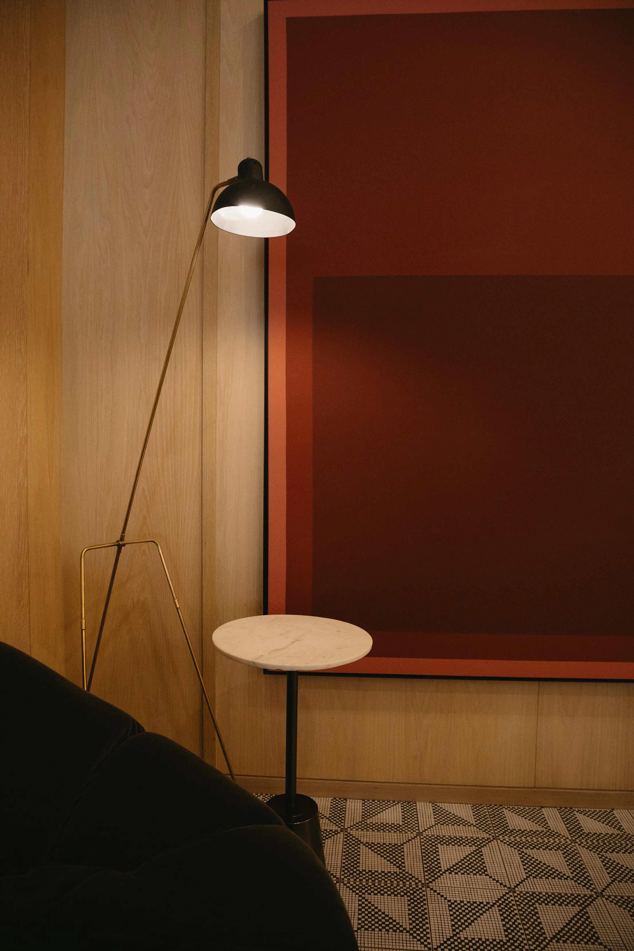 A contemporary interior scene with a black leather sofa, a tall floor lamp with a black shade, a small round marble-topped side table, wood-paneled walls, a large red and black geometric artwork, and a patterned tile floor.