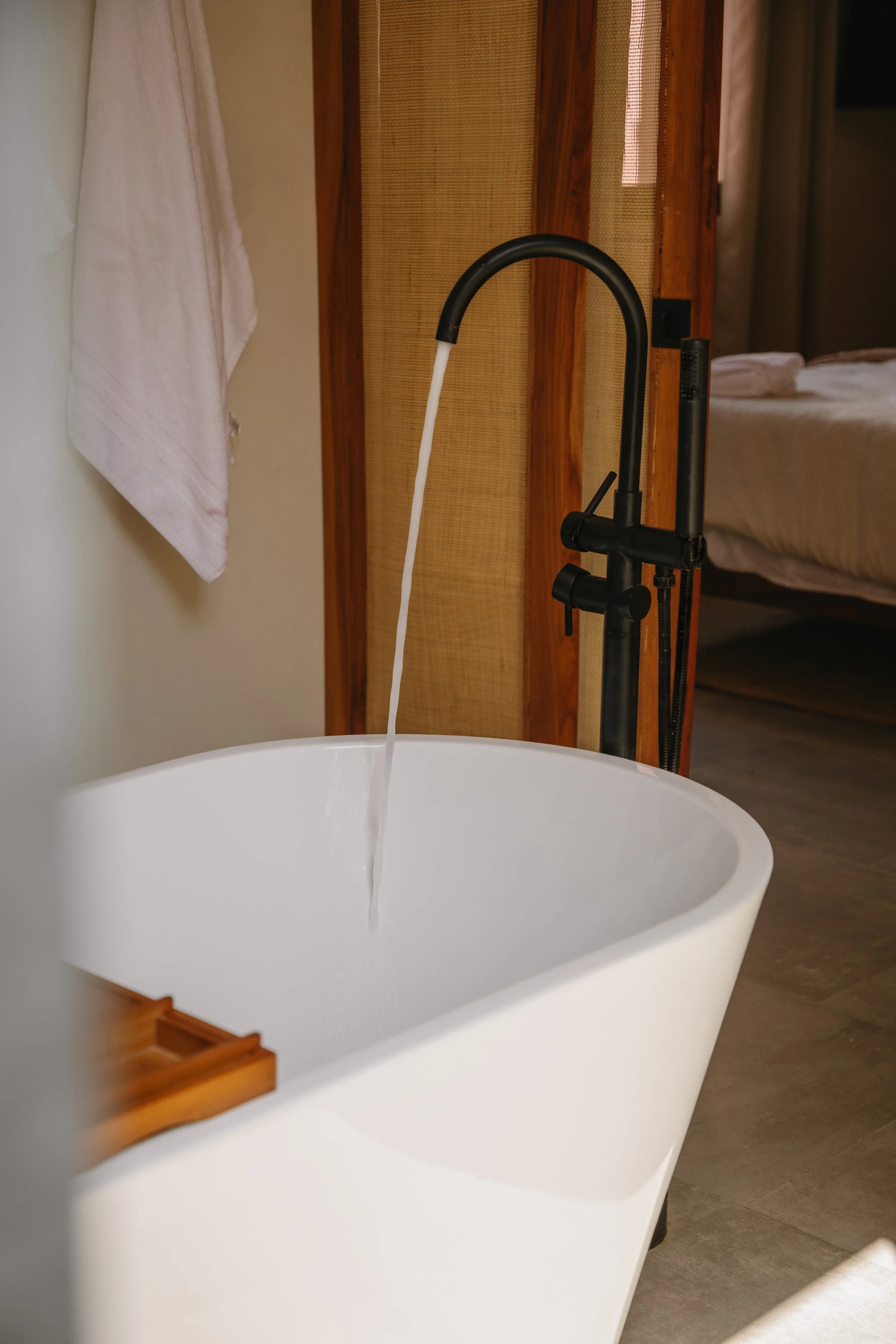 White freestanding bathtub in a bathroom with a black floor-mounted faucet, near a wooden privacy screen and a bed in the background