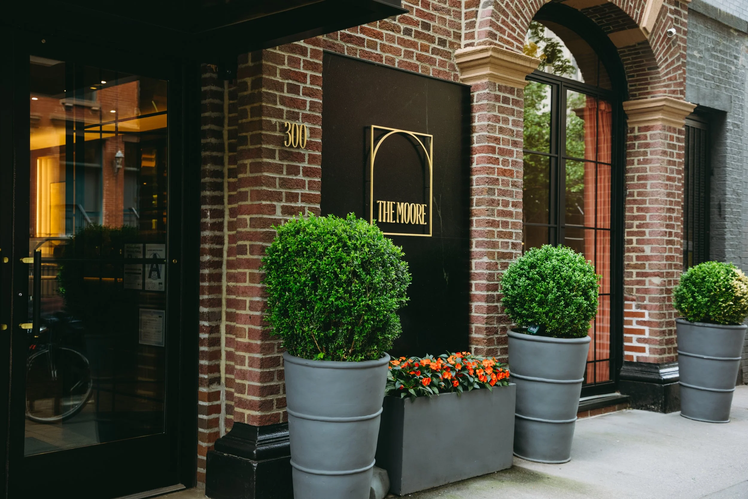 The exterior of a brick building with a black door, adorned with large potted green bushes and a rectangular planter with orange flowers. A sign reads 'The Moore' and the address number 300 is visible.