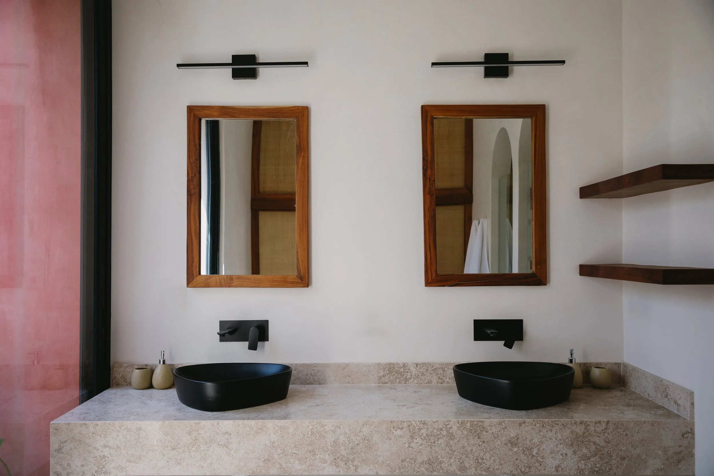 A modern double bathroom vanity with two black vessel sinks, two mirrors framed in wood, two wall-mounted black faucets, black light fixtures above each mirror, a speckled beige countertop, and two decorative vases on the left. There are two wooden s
