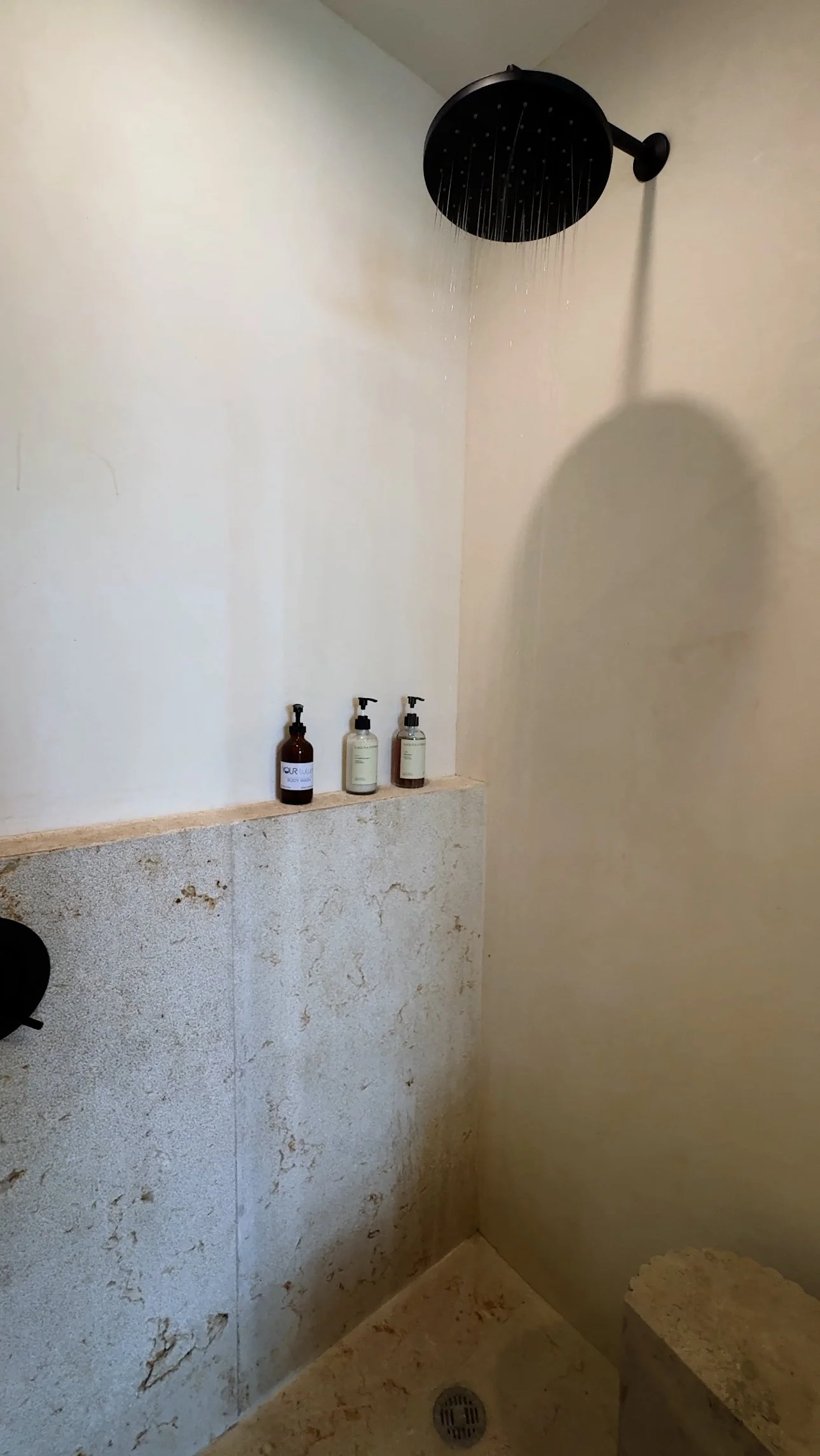 A shower area with a black rainfall showerhead attached to the ceiling. There are three bottles of bath products on a ledge, including a brown bottle and two clear bottles with black pump dispensers. The shower floor has a drain, and the wall below t