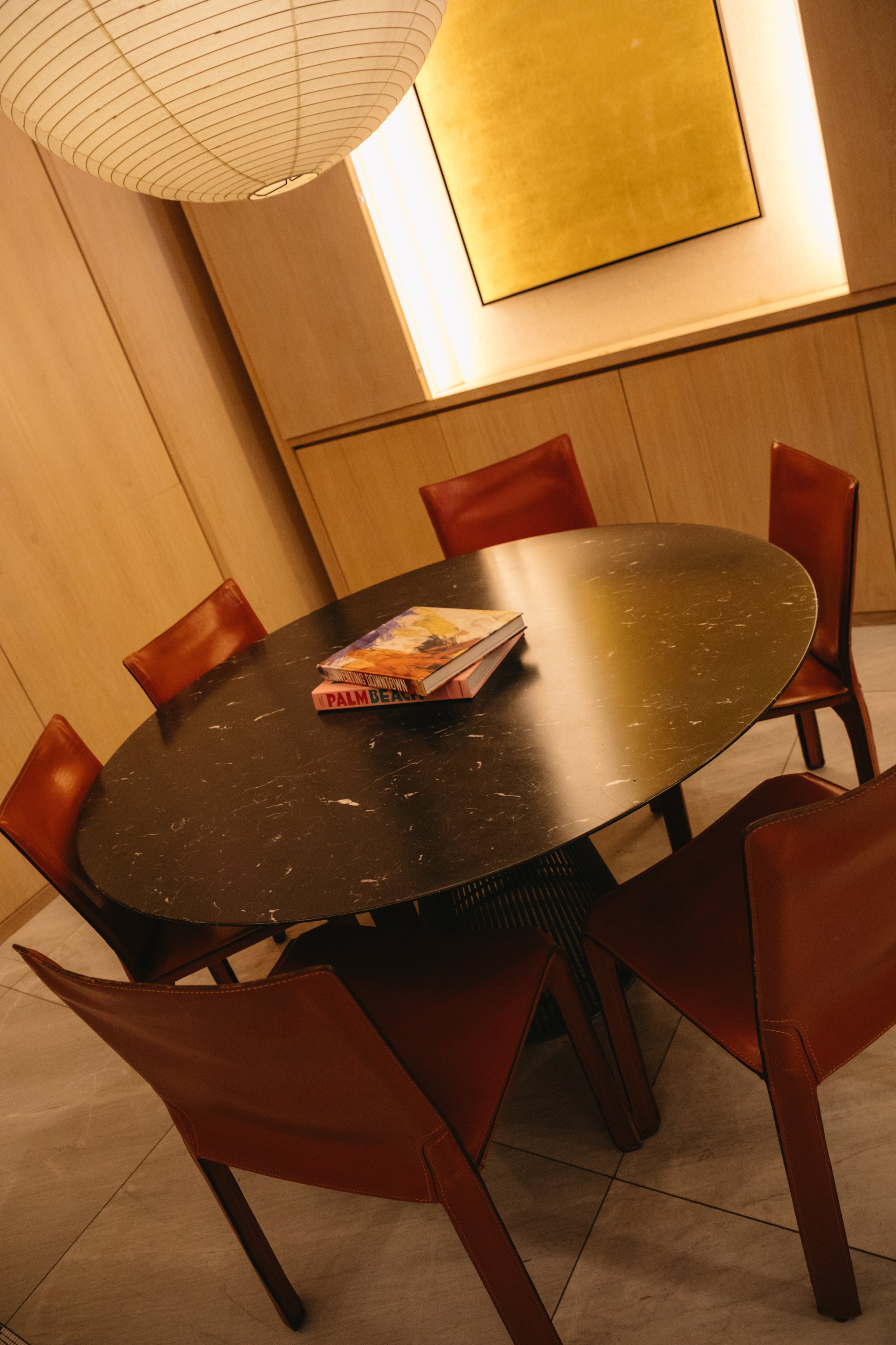 A round black marble table with six brown leather chairs surrounding it, in a warmly lit room with wooden wall panels and a yellow abstract art piece. There are four magazines on the table.