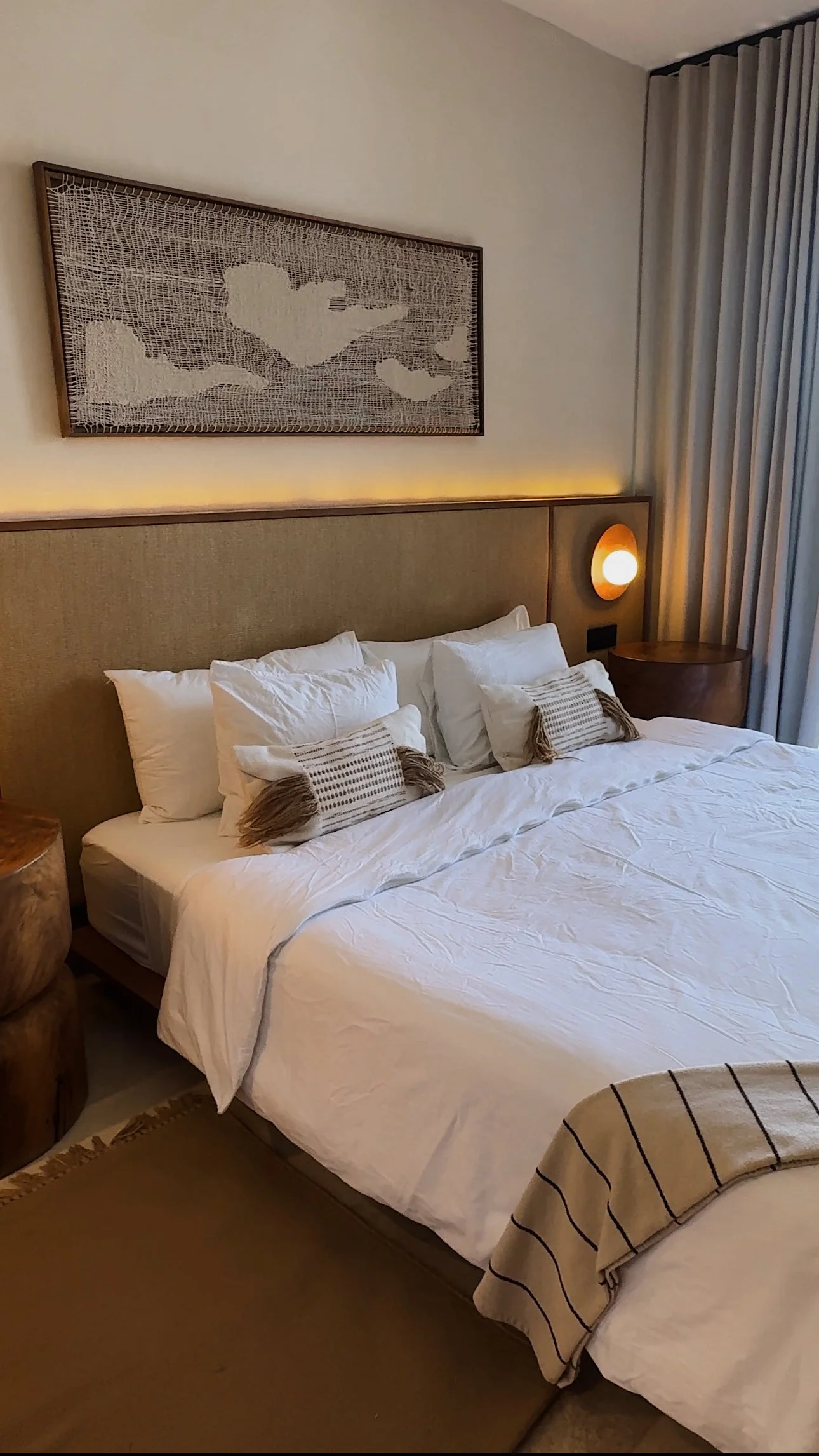 A neatly made bed with white linens and four pillows, two large and two decorative with tassels, in a modern hotel room with wooden nightstands and a textured wall art piece above the bed. A soft lighting feature runs along the headboard, and a wall-mounted round light is on the right side.
