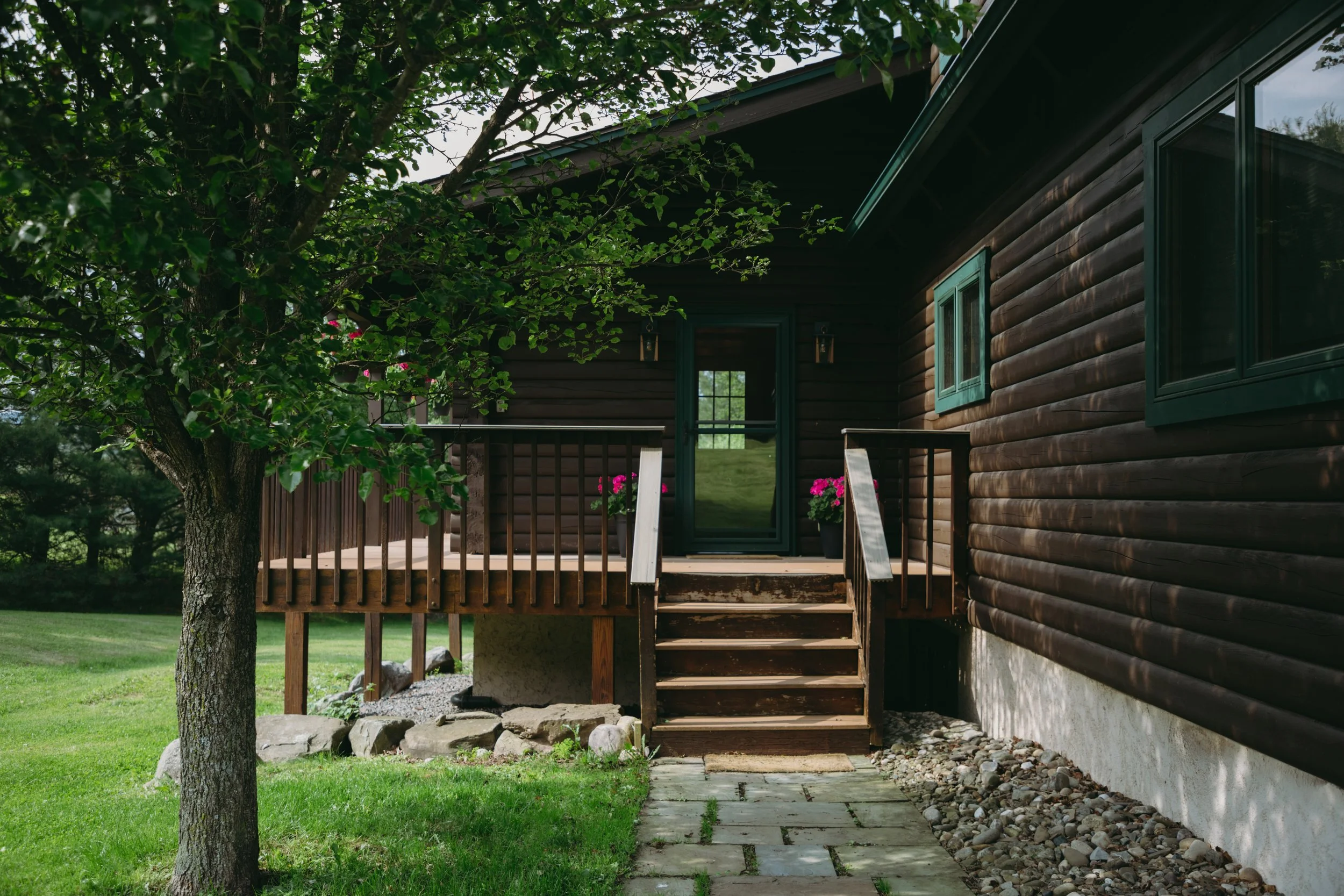 Wooden house with a small front porch, steps, potted pink flowers, a tree, and a stone pathway.