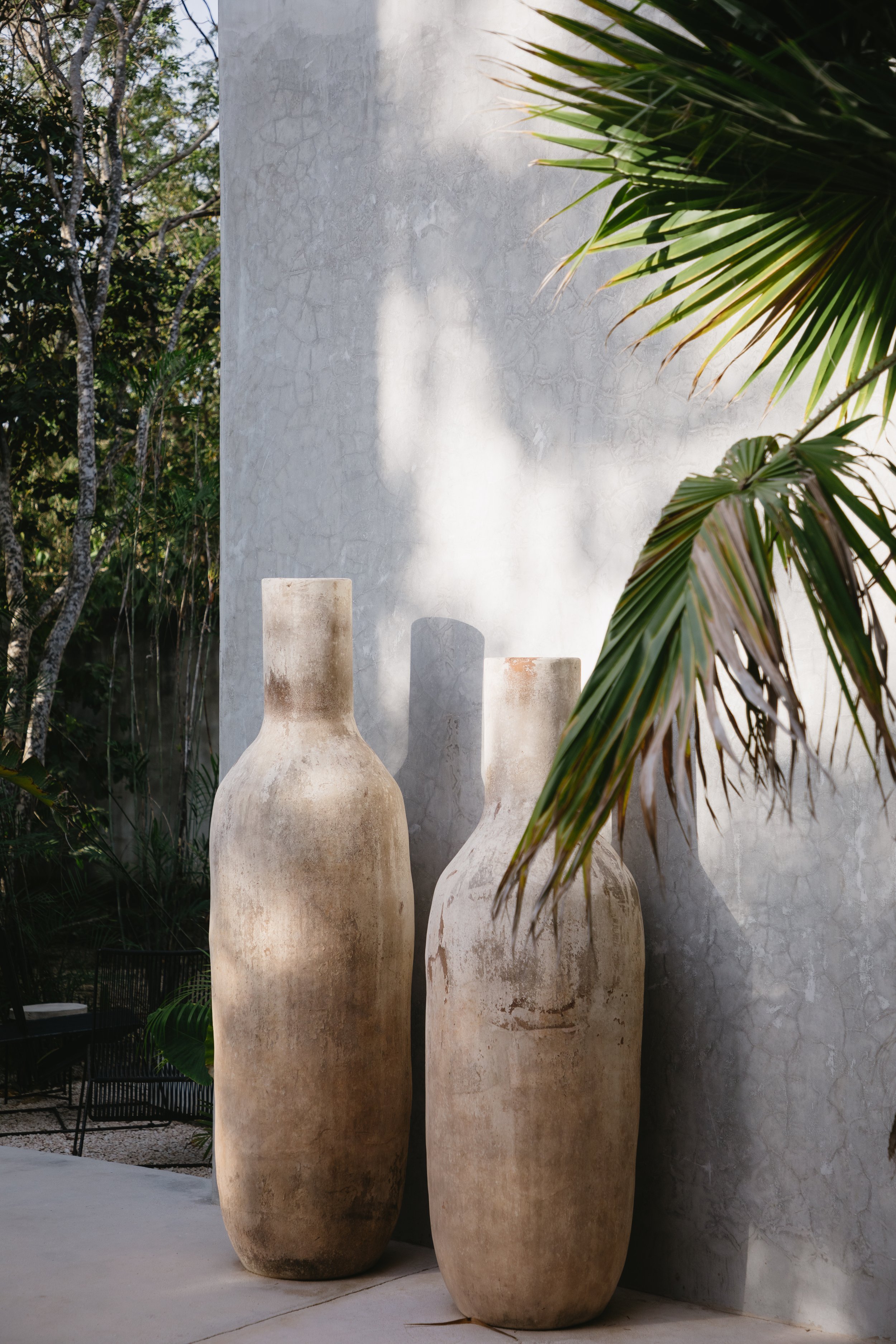 Three large, weathered clay vases placed outdoors beside a concrete wall, with dappled sunlight and green foliage nearby.