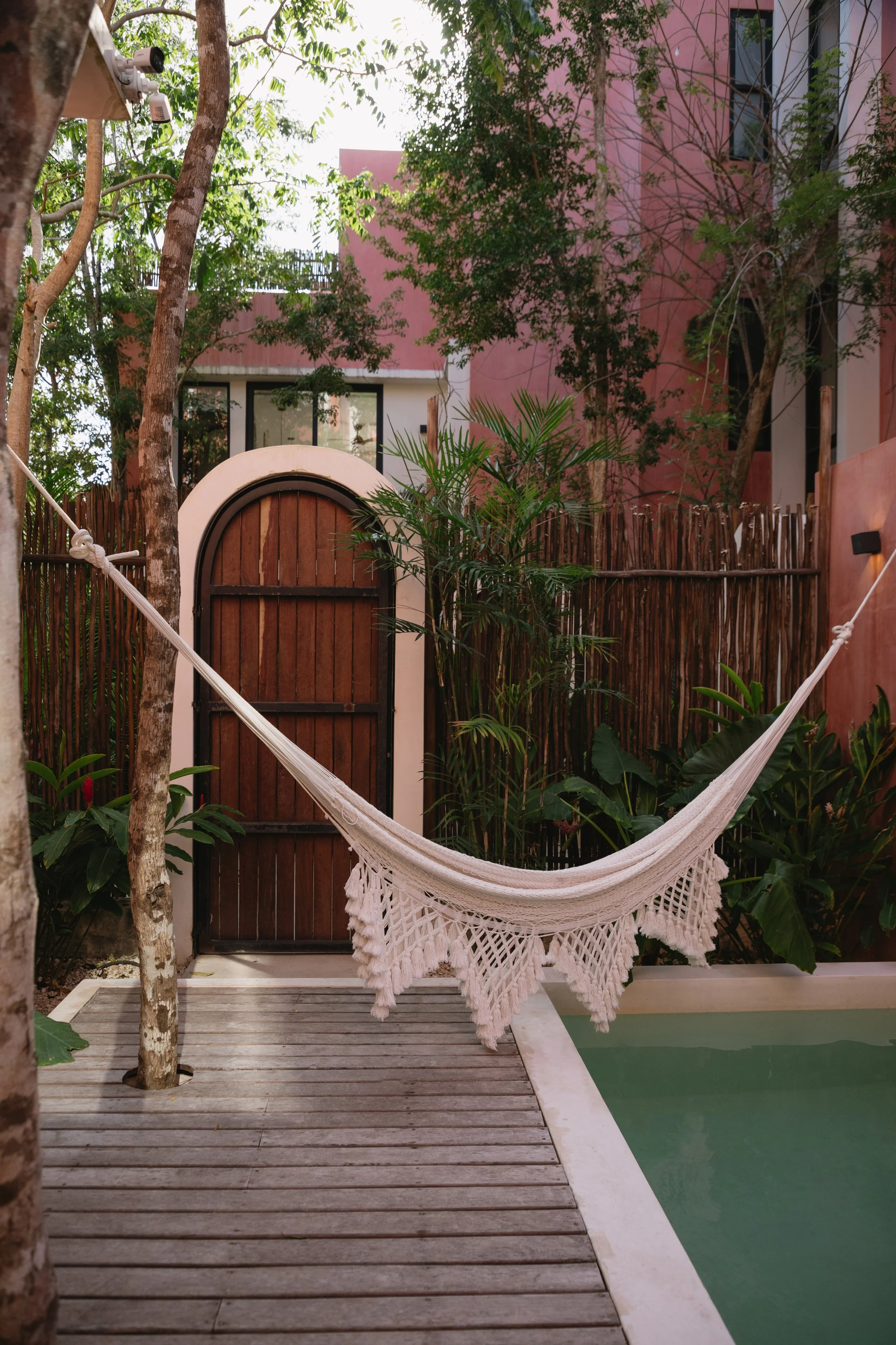 A white hammock with decorative tassels hanging between trees in an outdoor garden area with a wooden deck and a pool, surrounded by lush plants and a pink building in the background.