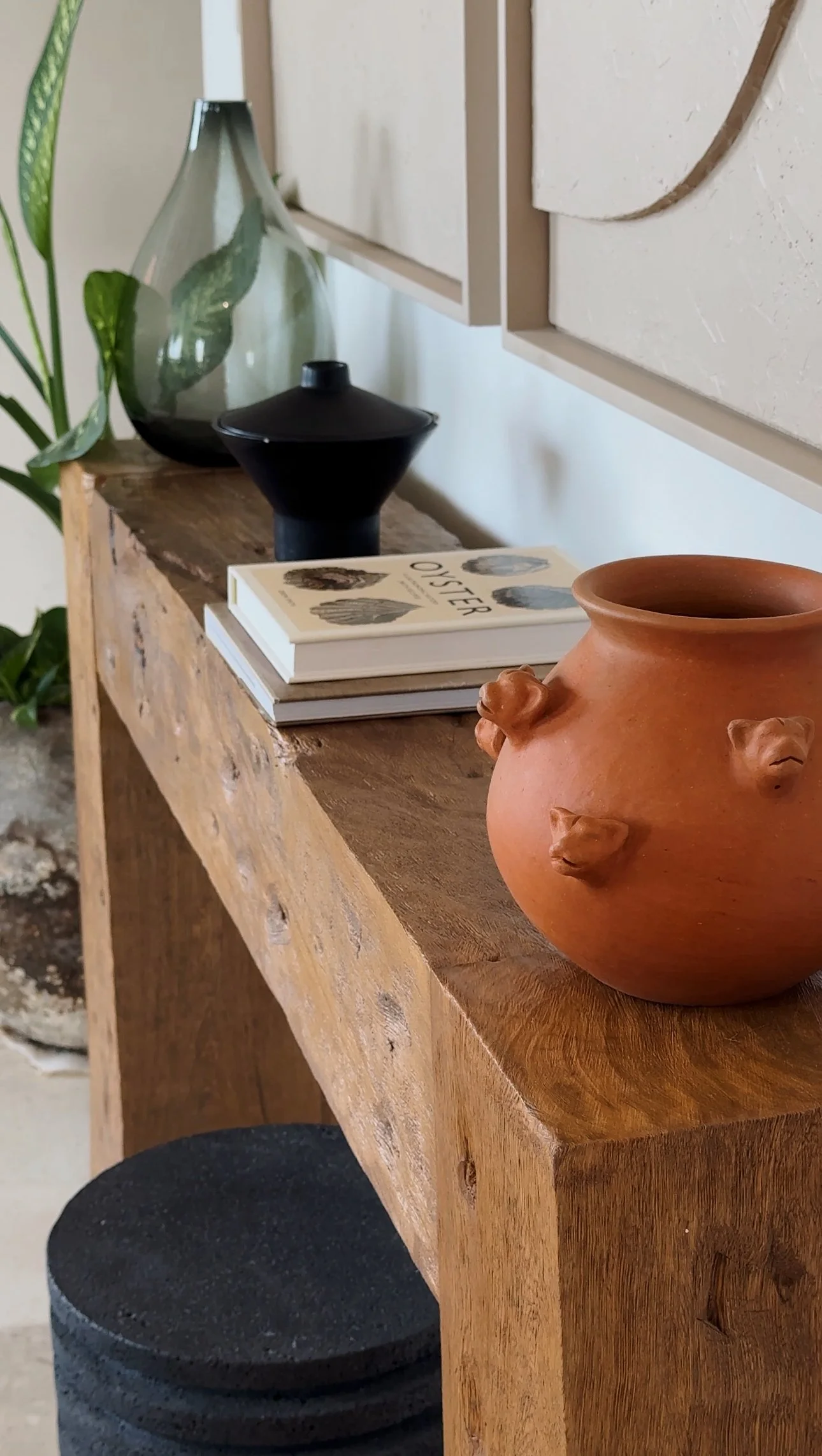 A wooden console table decorated with a terracotta vase, stacked books, a black decorative object, and a green glass vase, with a plant nearby.