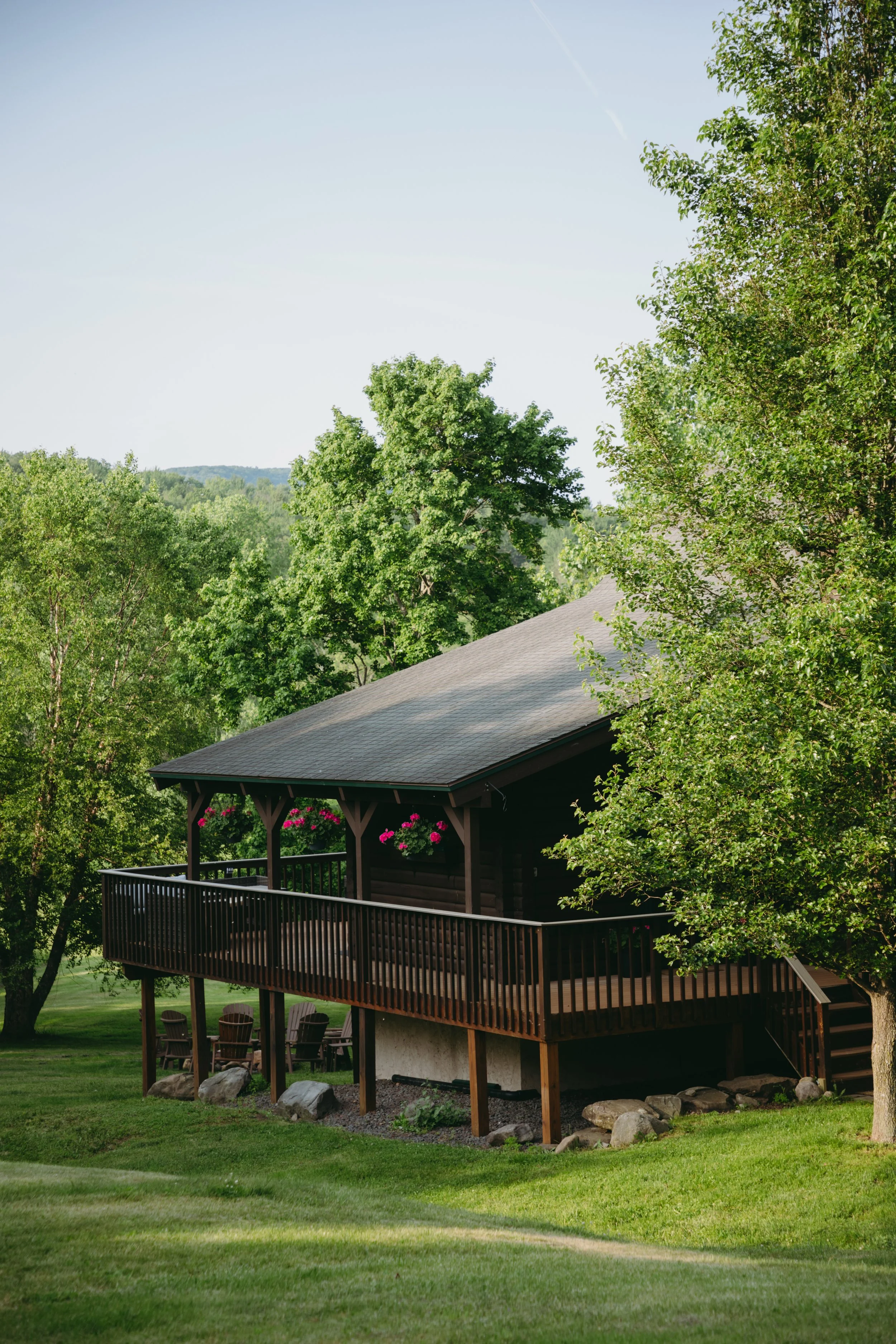 A wooden house with a large deck surrounded by green trees and grass, under a clear blue sky.