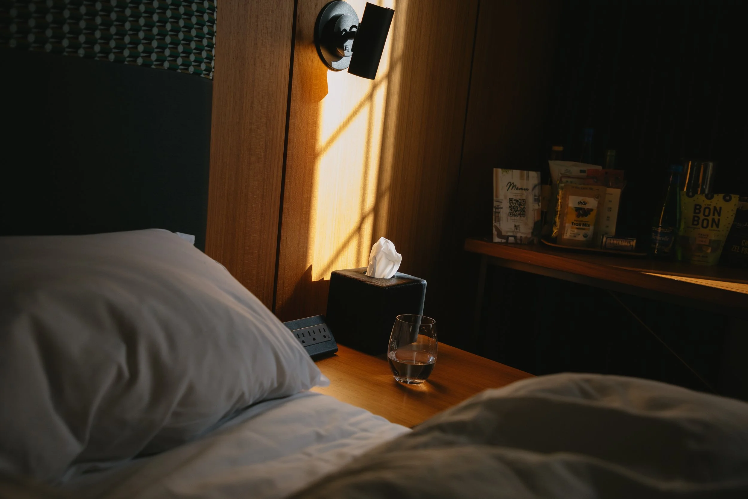 A hotel room with a bed, a nightstand with a glass of water, a tissue box, and a wall-mounted lamp, with sunlight casting shadows on the wooden wall and ceiling.