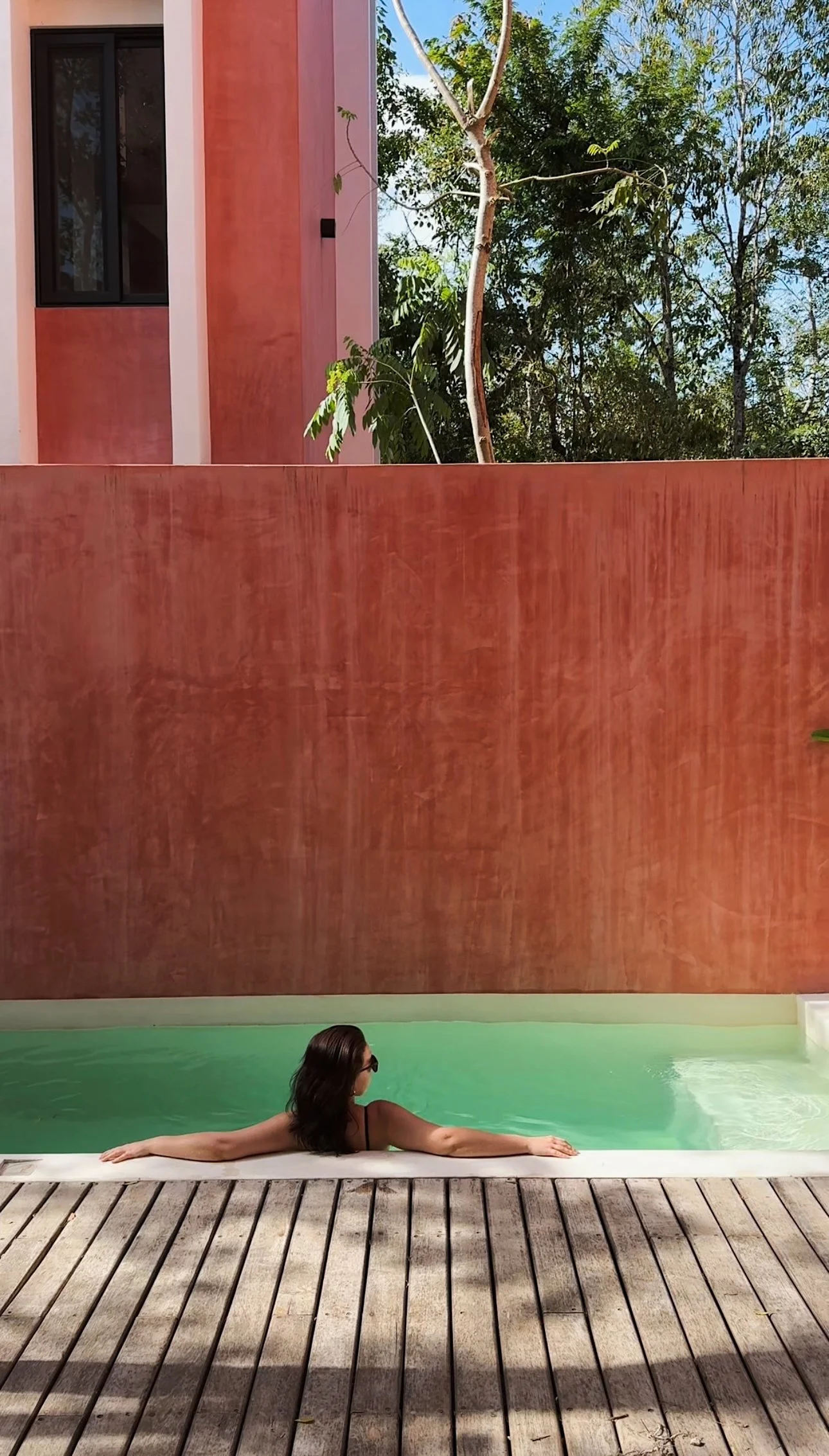 Woman lounging in a swimming pool with a wooden deck in the foreground, a tall brick wall behind her, and trees with blue sky above.
