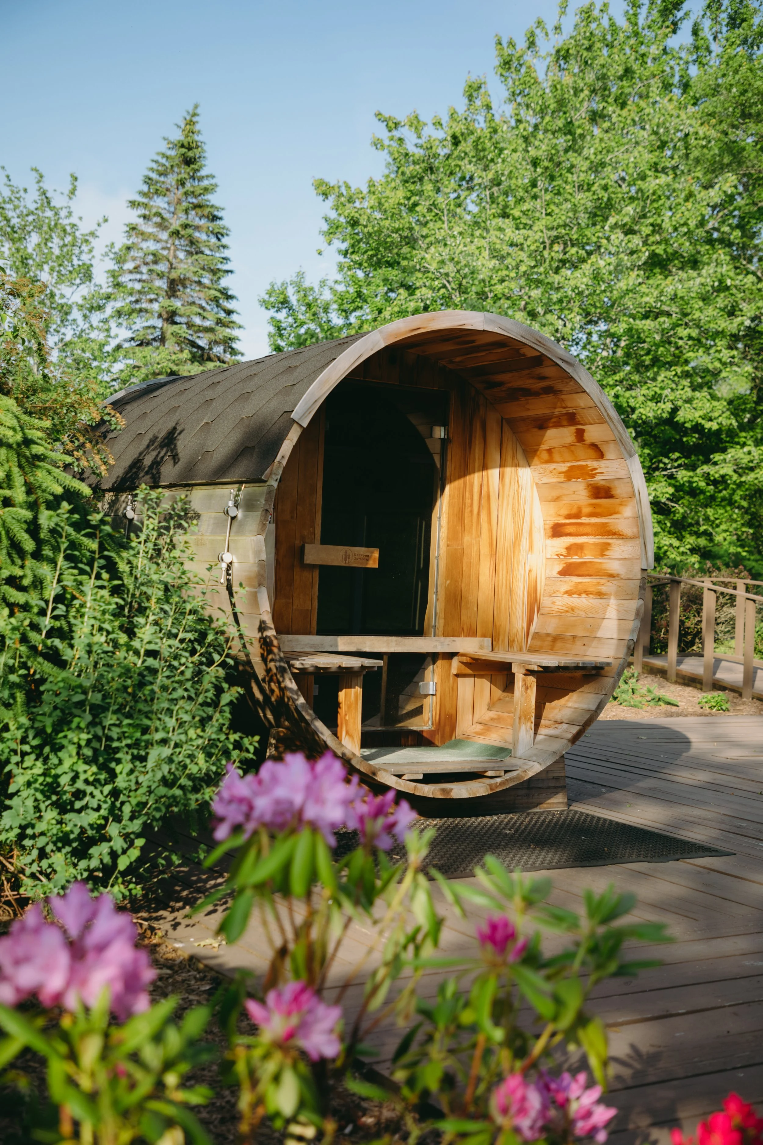 A wooden sauna structure shaped like a large barrel with a rounded entrance, situated outdoors on a wooden deck surrounded by greenery and pink flowers, under a clear blue sky.