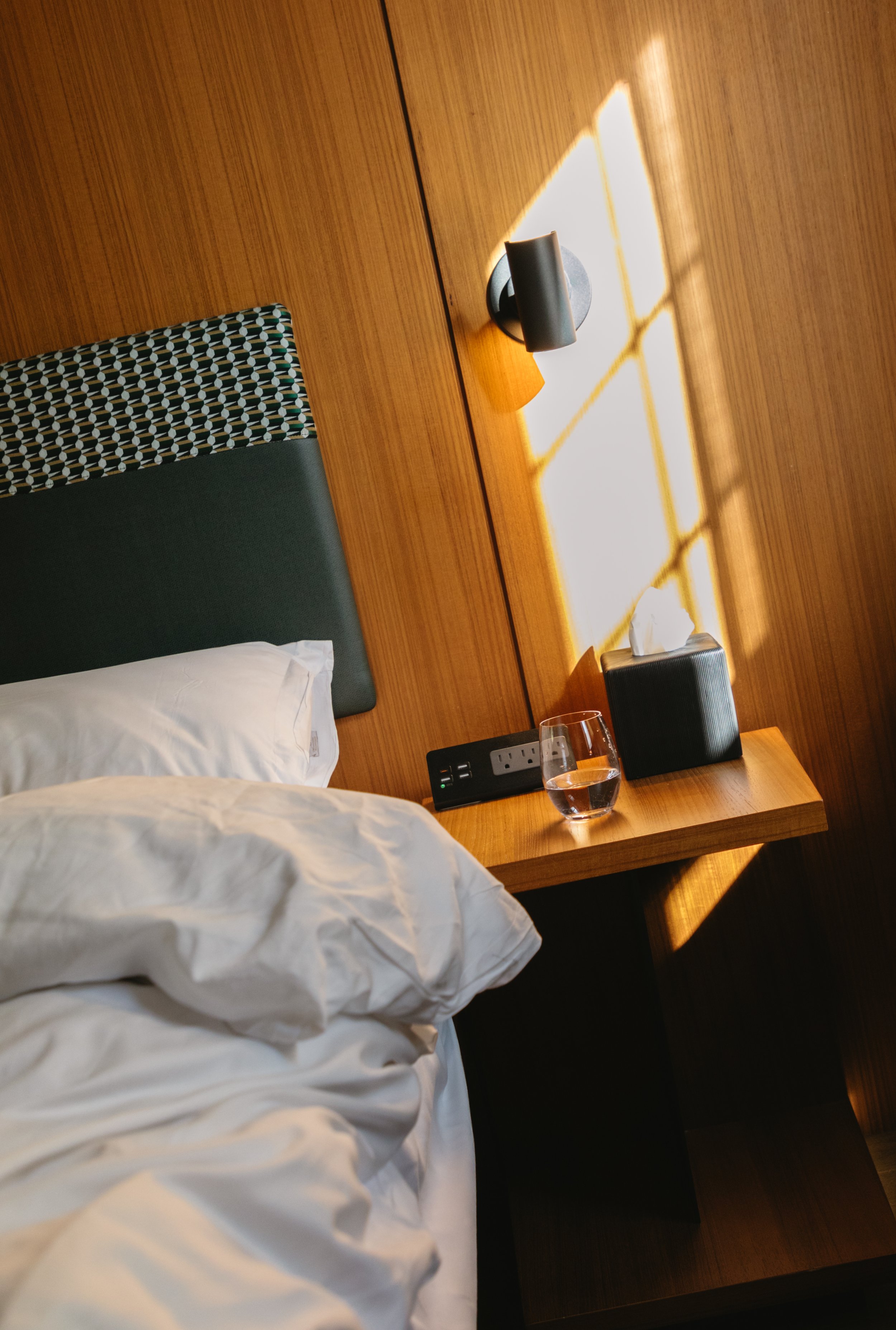 A bedroom with a bed, a wooden headboard, a nightstand with a glass of water, a tissue box, a power outlet, and a wall sconce. Sunlight casts a grid pattern on the wall.