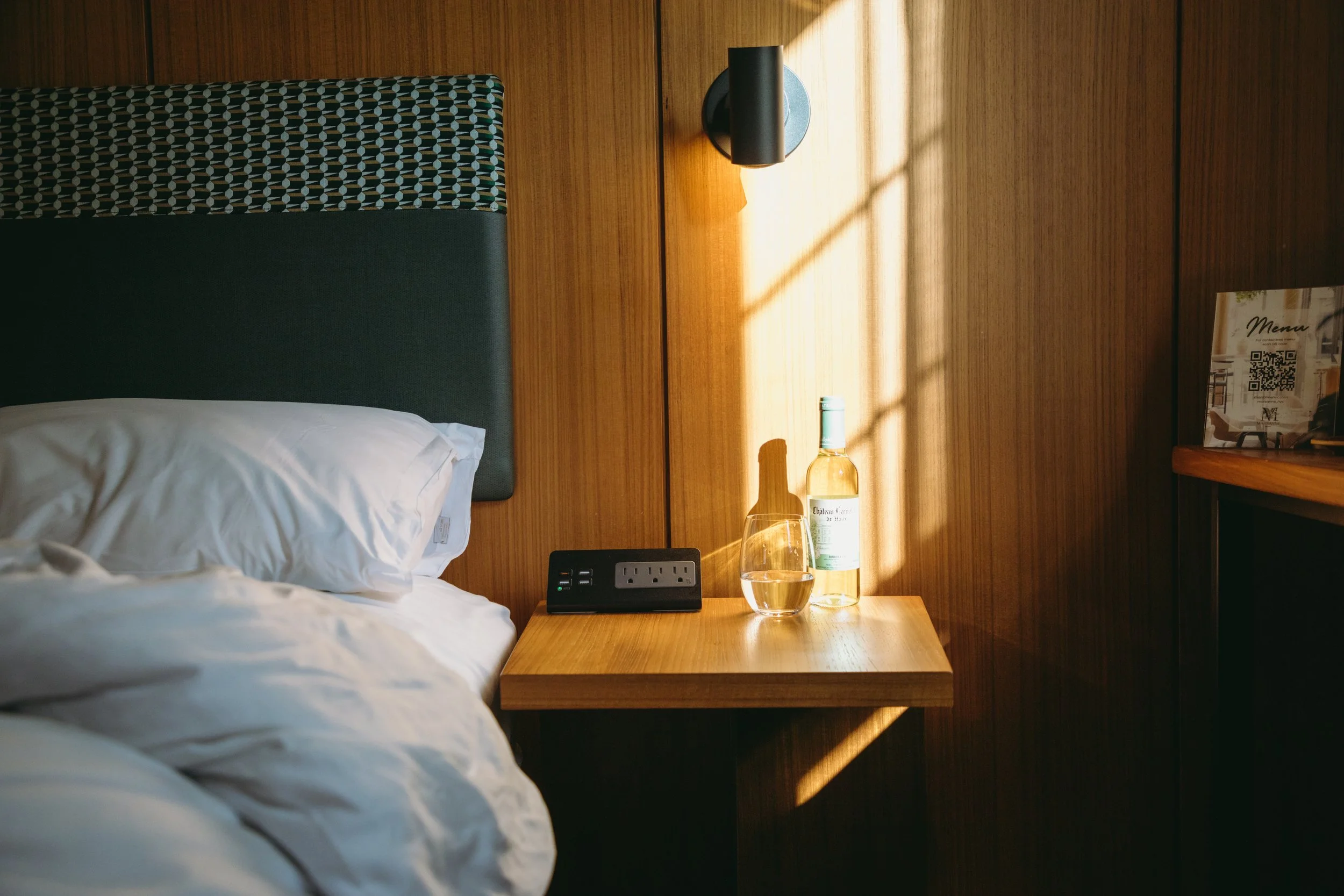 A bedside table with a bottle of water, a glass filled with water, a power strip with plugs, and a digital clock, next to a bed with white rumpled sheets and pillows, against a wooden wall with a wall-mounted lamp casting light and shadow.