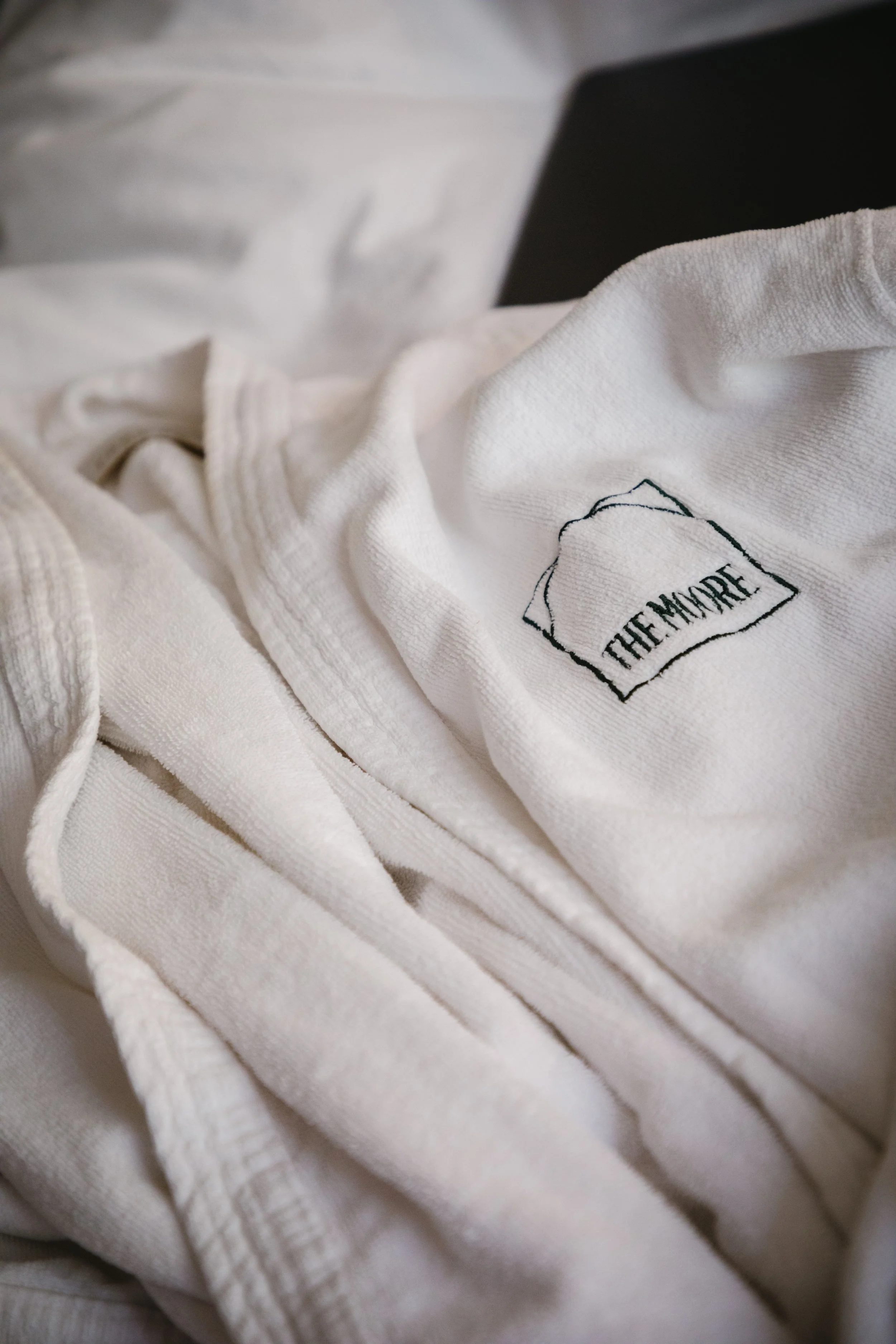 Close-up of white embroidered clothing, possibly a kimono or jacket, with a logo that reads 'THE MADE' inside a rough outlined rectangle.