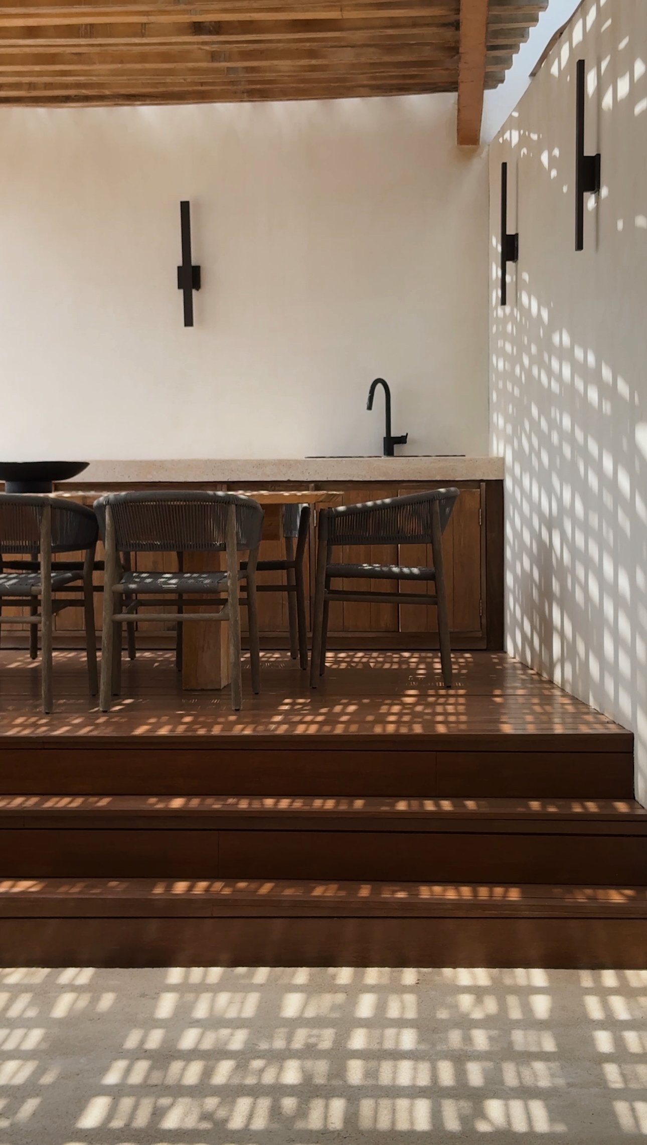 Outdoor patio with wooden flooring and stairs, featuring a table and chairs, a white wall with black wall-mounted lights, and a window or opening casting patterned shadows.
