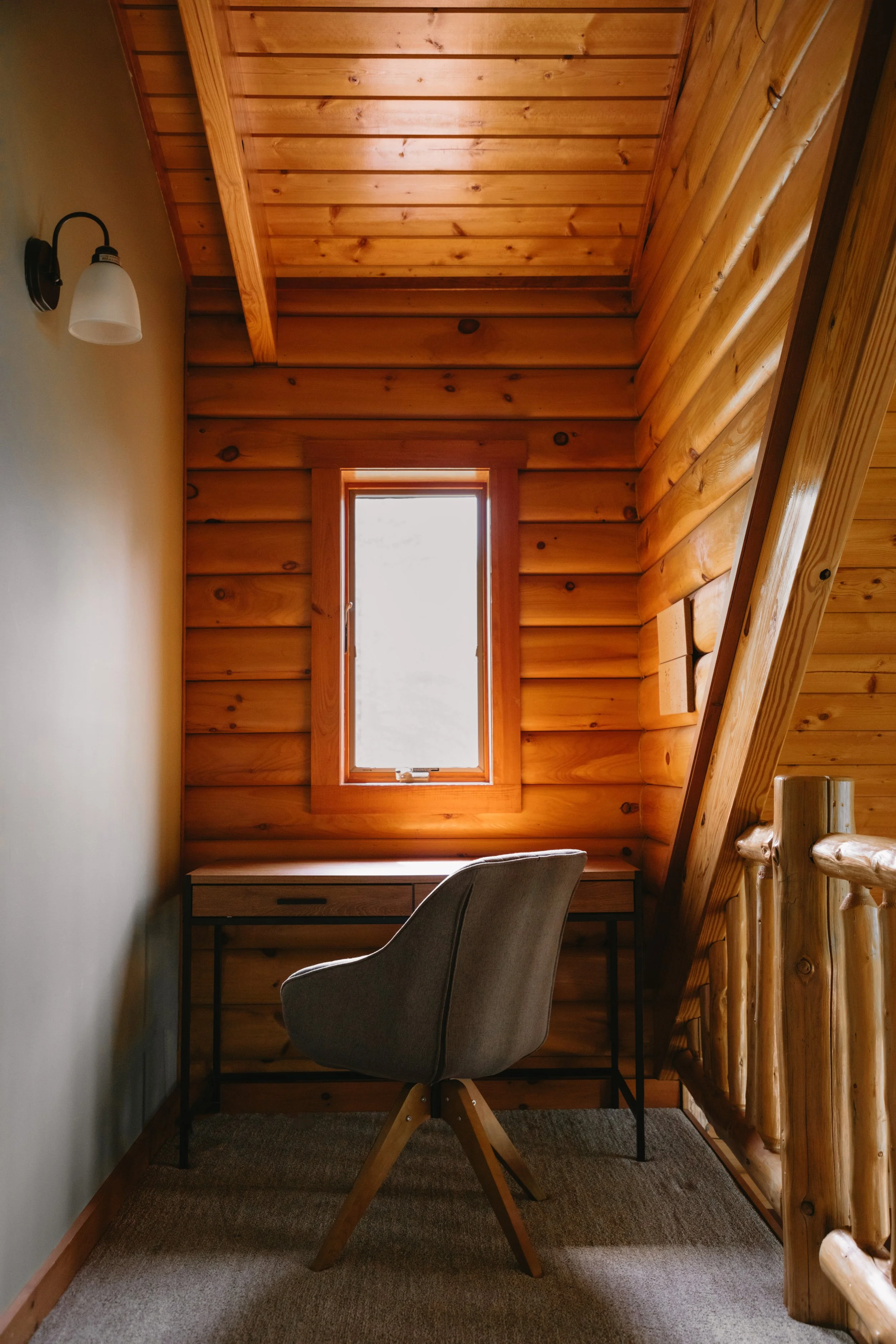 A small wooden room with a slanted ceiling, a window, a desk, and an upholstered chair.