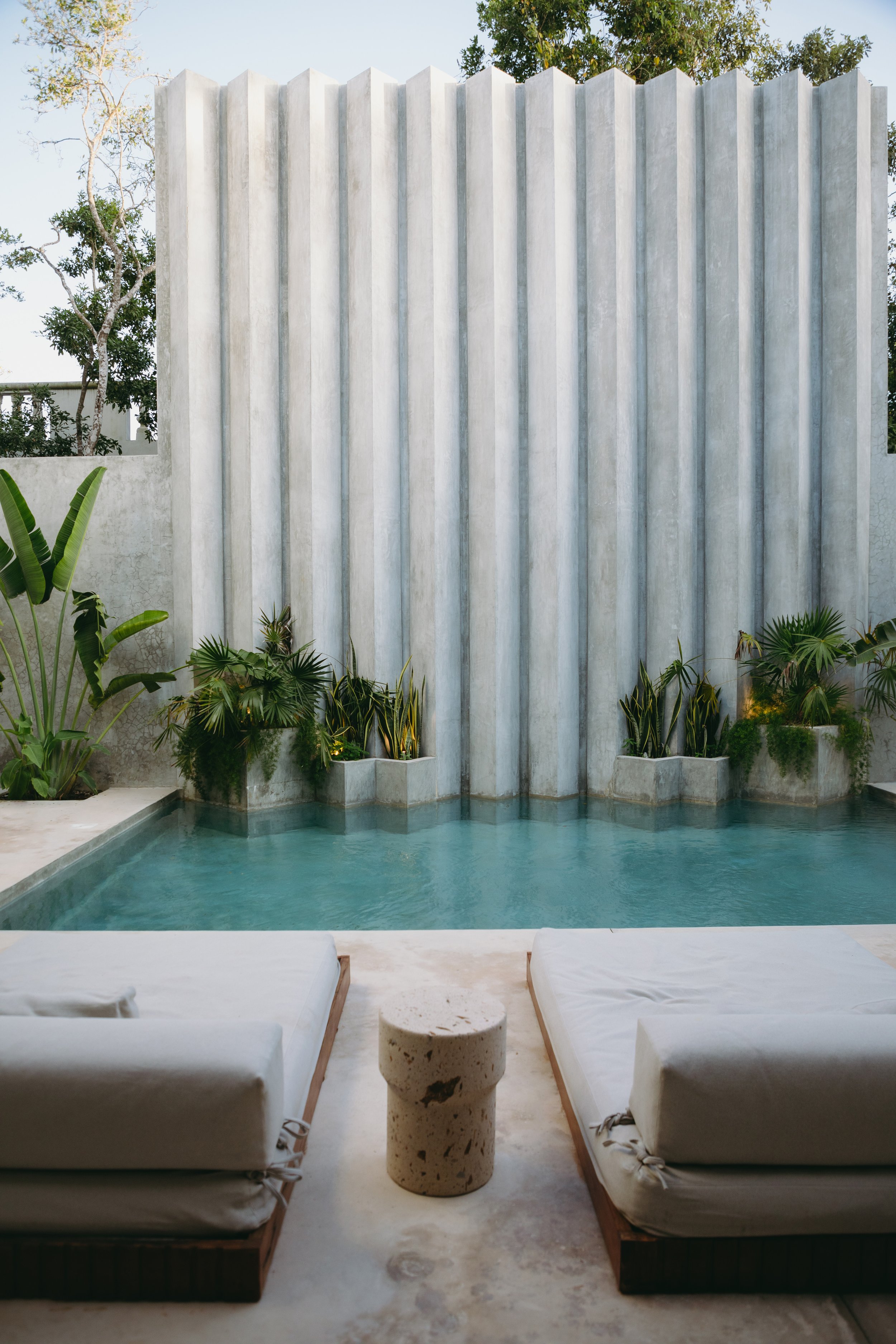A luxurious outdoor pool with a waterfall feature, surrounded by tropical plants, with two lounge chairs and a small round table in the foreground.