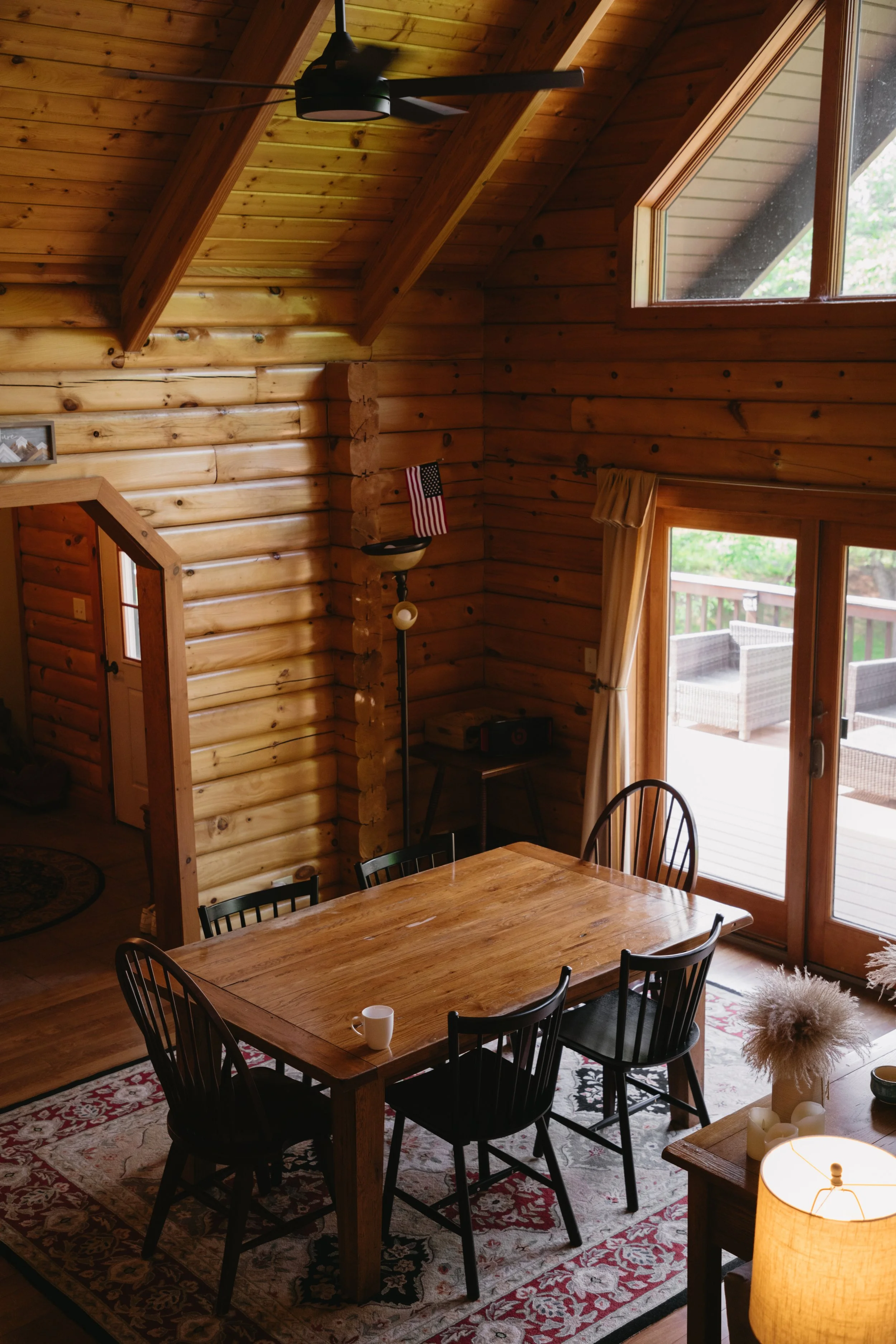 The interior of a log cabin dining room featuring a wooden table with four black chairs, a patterned area rug, a sliding glass door leading to an outdoor deck with wicker furniture, and large windows allowing natural light.