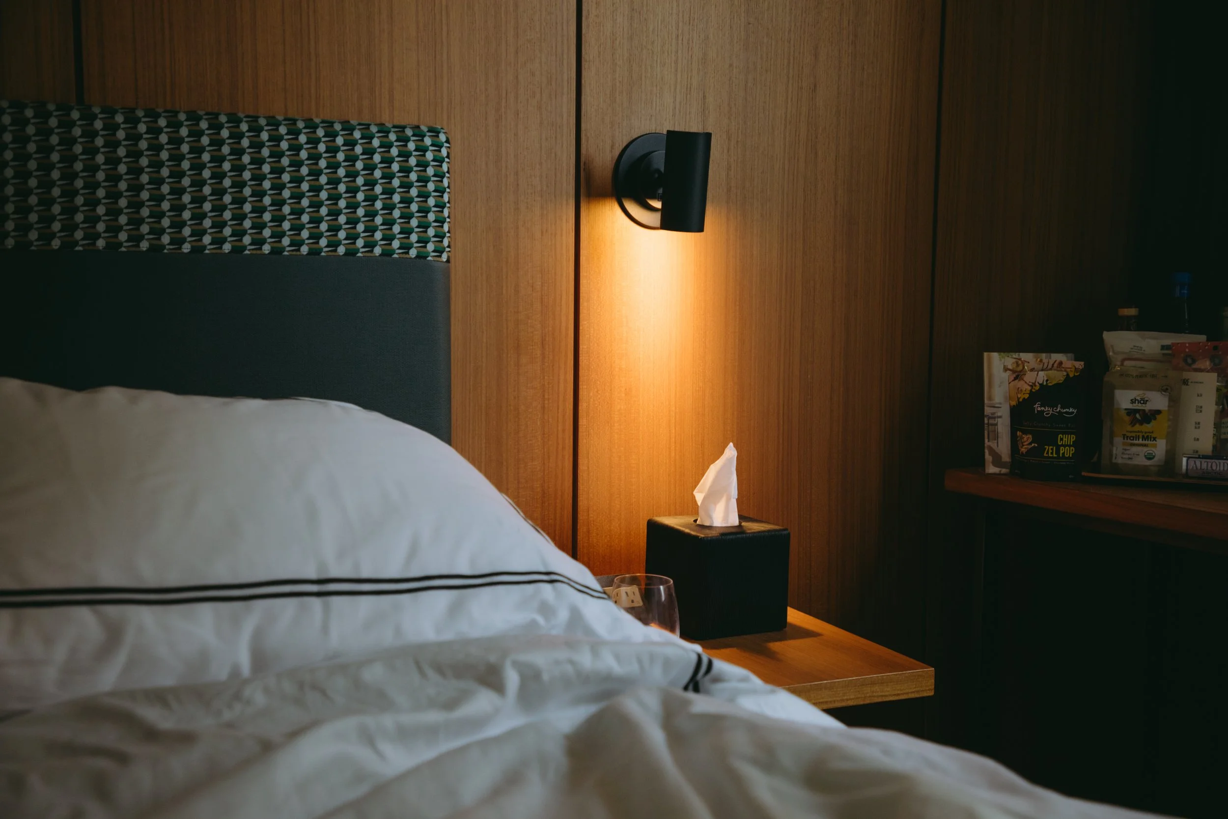 A cozy hotel bedroom with a bed, bedside table, tissues, a wall-mounted black reading light, and snacks on a shelf.