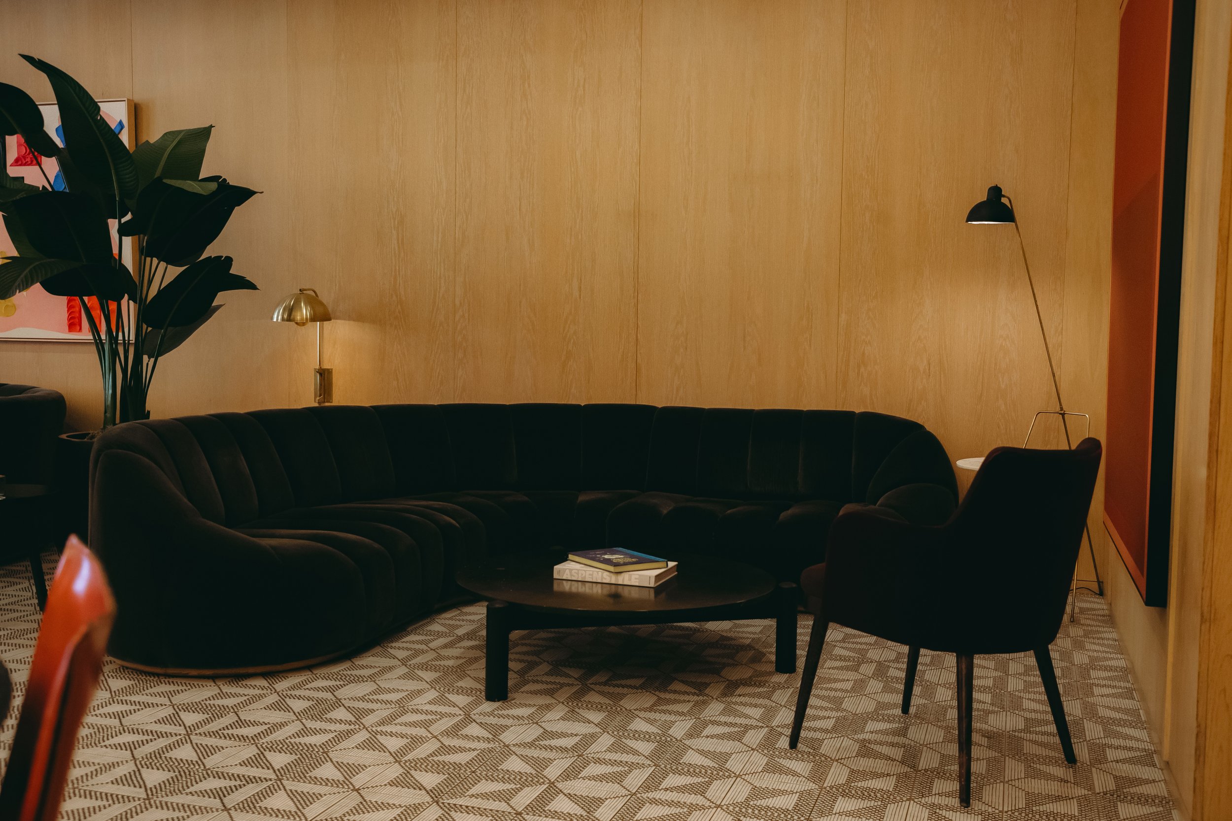 A cozy living room with a dark green velvet sectional sofa, a round black coffee table with books, a single dark upholstered armchair, a large potted plant, and modern wall lamps, all against warm wooden wall panels.