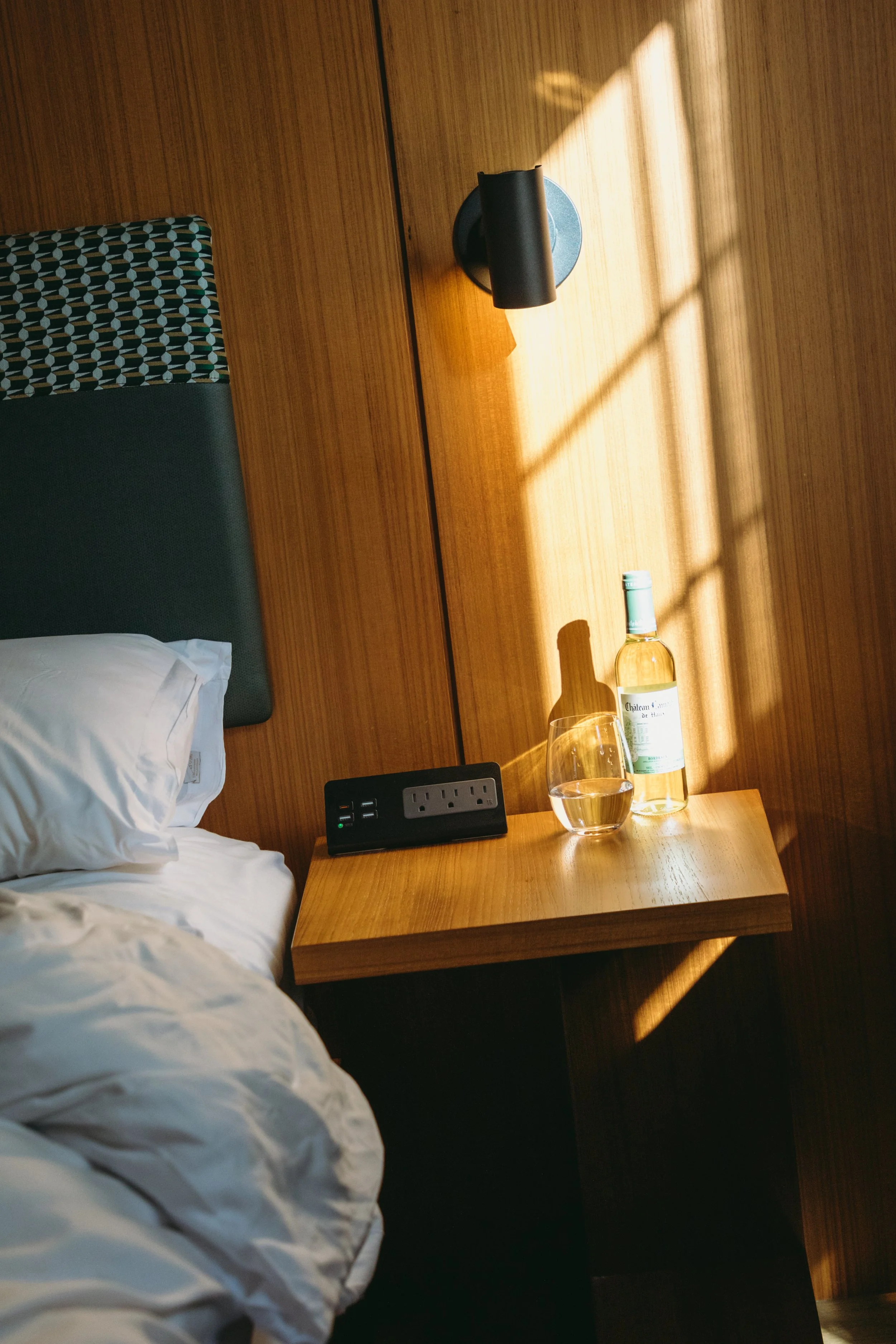 A bedside table with a bottle of white wine, a glass of wine, an electrical outlet, and a wall-mounted lamp, with sunlight casting shadows on the wall.